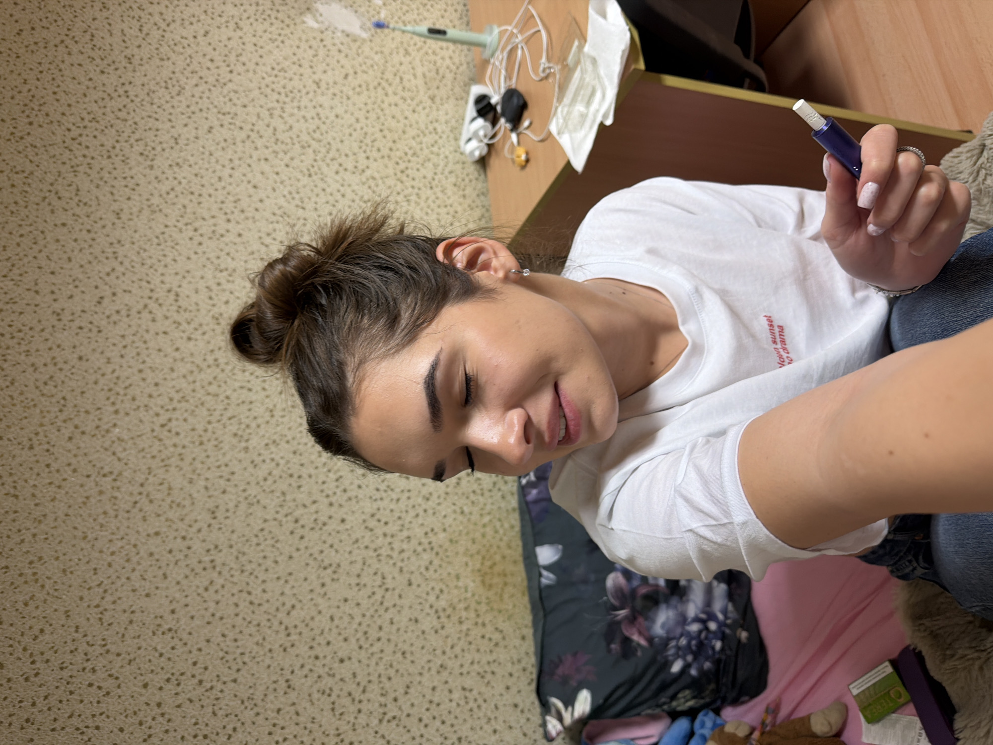 A woman is holding an IQOS device, a type of heat-not-burn tobacco product. The location is inside a building in Sofia, Bulgaria, based on the presence of an IQOS device which are commonly used in Bulgaria. The woman has brown hair styled in a bun, a white t-shirt, and blue jeans. She is holding the IQOS device in her right hand. The environment includes a desk with electronic items, floral bedding, and carpeting.