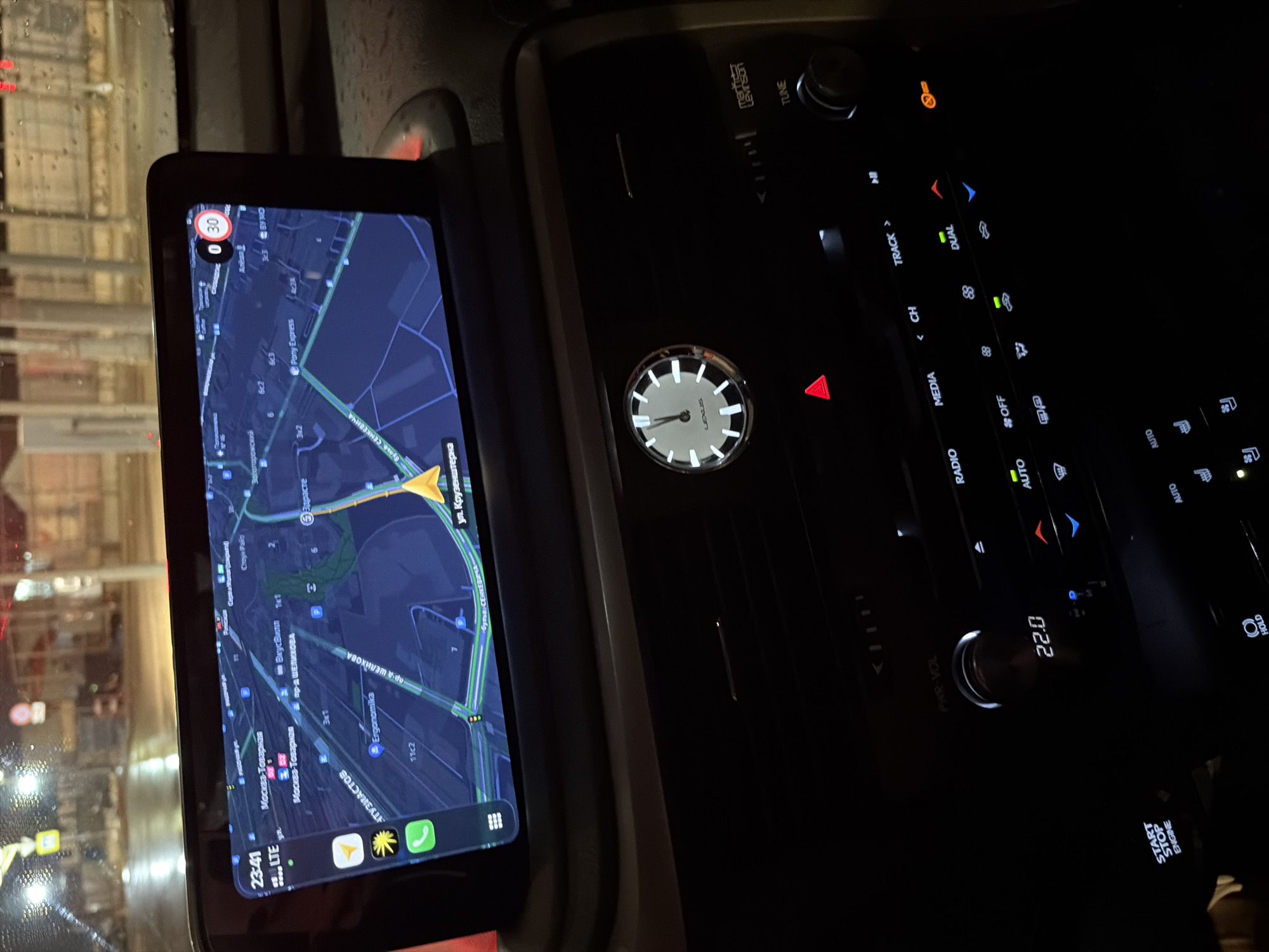 The image shows the interior of a Lexus car at night, likely in Moscow, Russia. A navigation system is displayed on the car's screen, showing a map with routes and street names. The navigation shows "ул. Крузенштерна" (Kruzenstern Street) and "Москва-Товарная" (Moscow-Tovarnaya). The car's climate control panel is visible below the screen, displaying the temperature at 22.0 degrees Celsius. A clock with Roman numerals is also integrated into the dashboard. The ambient lighting is low, suggesting nighttime.