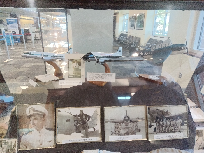 The image displays an aviation historical exhibit housed within a glass display case inside an airport terminal in the city of Park Hill, United States. The display case contains three model airplanes and four framed photographs. From left to right, the model aircraft include a white commercial passenger jet with green stripes, a white multi-engine propeller airliner with dark accents, and a dark military propeller aircraft marked with the number '61' on its tail and wing. All models are mounted on wooden stands. Small informational plaques with text are situated near the model bases. Below the models, resting on a dark surface, are four framed photographs. The leftmost is a color portrait of a man in a white naval officer's uniform. The subsequent three are black and white photographs: one depicts an individual in flight gear next to a military aircraft marked '35'; another shows a military jet aircraft viewed from the front with ground crew; and the last features a group of uniformed personnel posing in front of a military jet. Reflections on the display glass reveal parts of the surrounding airport environment, including a 'TSA Pre✓®' sign and queue barriers on the left, and rows of grey airport seating, large windows, and framed artwork on the right.