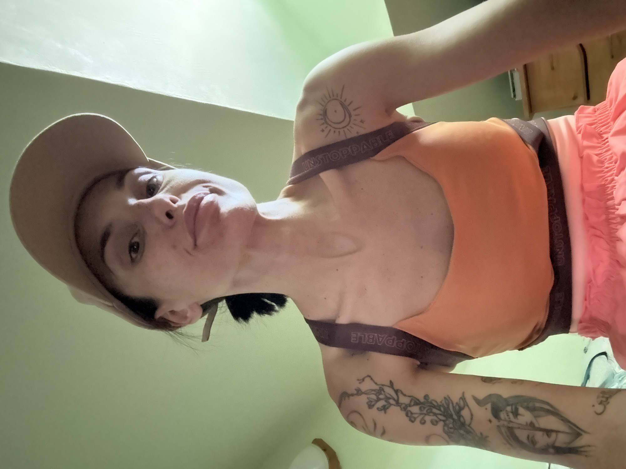 An indoor, low-angle, medium shot captures a person with dark hair, wearing a tan baseball cap, an orange sports bra, and pink shorts. The person's face is partially visible, turned towards the camera, with a neutral expression. Tattoos are visible on their left arm and shoulder, including a floral design and a depiction of a face. The background consists of a light green wall and a wooden dresser. The image appears to be a selfie. There is no indication of the city of Pleven, Bulgaria in the image.