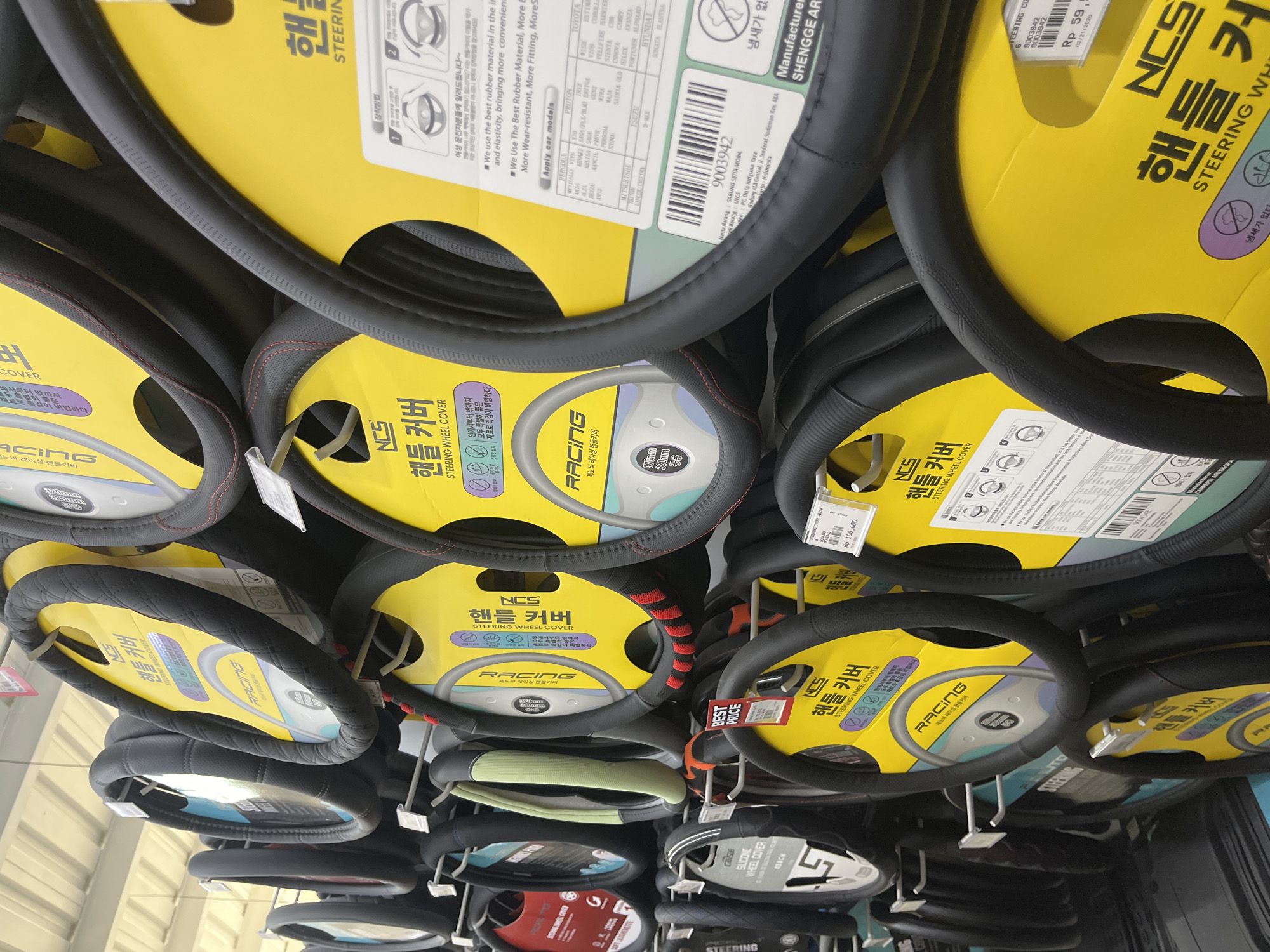 This image captures a retail display of numerous steering wheel covers, likely in an auto parts store or a section of a general merchandise store in West Jakarta, Indonesia. The covers are packaged in bright yellow and white/blue cardboard with clear plastic rings, and they are hanging vertically from metal hooks on a rack.
The primary subjects are the steering wheel covers themselves, presented in various designs. Most are black, with some featuring red stitching, patterned textures, or light green accents. The packaging prominently displays the brand "NCS 핸들커버" (NCS Steering Wheel Cover) and "RACING."
Visible text provides detailed information about the products. The packaging includes instructions for installation and highlights features such as "best rubber material," "wear-resistant," "more fitting," and "냄새가 없다" (no smell). Compatibility lists numerous car models from manufacturers like PERODUA, PROTON, TOYOTA, MITSUBISHI, ISUZU, and HYUNDAI. Specific dimensions like "370mm" and "380mm" are also noted. One package mentions "SOFT LEATHERETTE" and another "SILICONE WHEEL COVER."
Price tags indicate costs in Indonesian Rupiah, such as "Rp 59,xxx" and "Rp 100,000," with one tag featuring "BEST PRICE." Manufacturer information points to "SHENGGE AR" (Shengge Auto Rubber), and distributor details show "PT. Duta Intiguna Yasa" located in "Jakarta-Indonesia," confirming the provided location context. A date "02/21/2026" is also visible, possibly an expiration or production date. The overall scene depicts a well-organized retail environment showcasing a variety of automotive accessories.