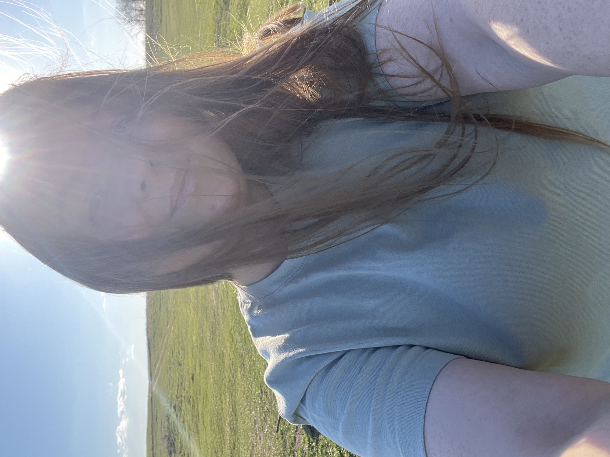 A selfie taken in an outdoor, grassy field setting. The subject appears to be a fair-skinned individual with reddish-brown hair. The sun is shining brightly, causing glare on the lens, obscuring the subject's facial features. The subject is wearing a light-colored, possibly green or blue, t-shirt. The background is a green field under a clear, blue sky. The location is possibly Раздолье, Russia.