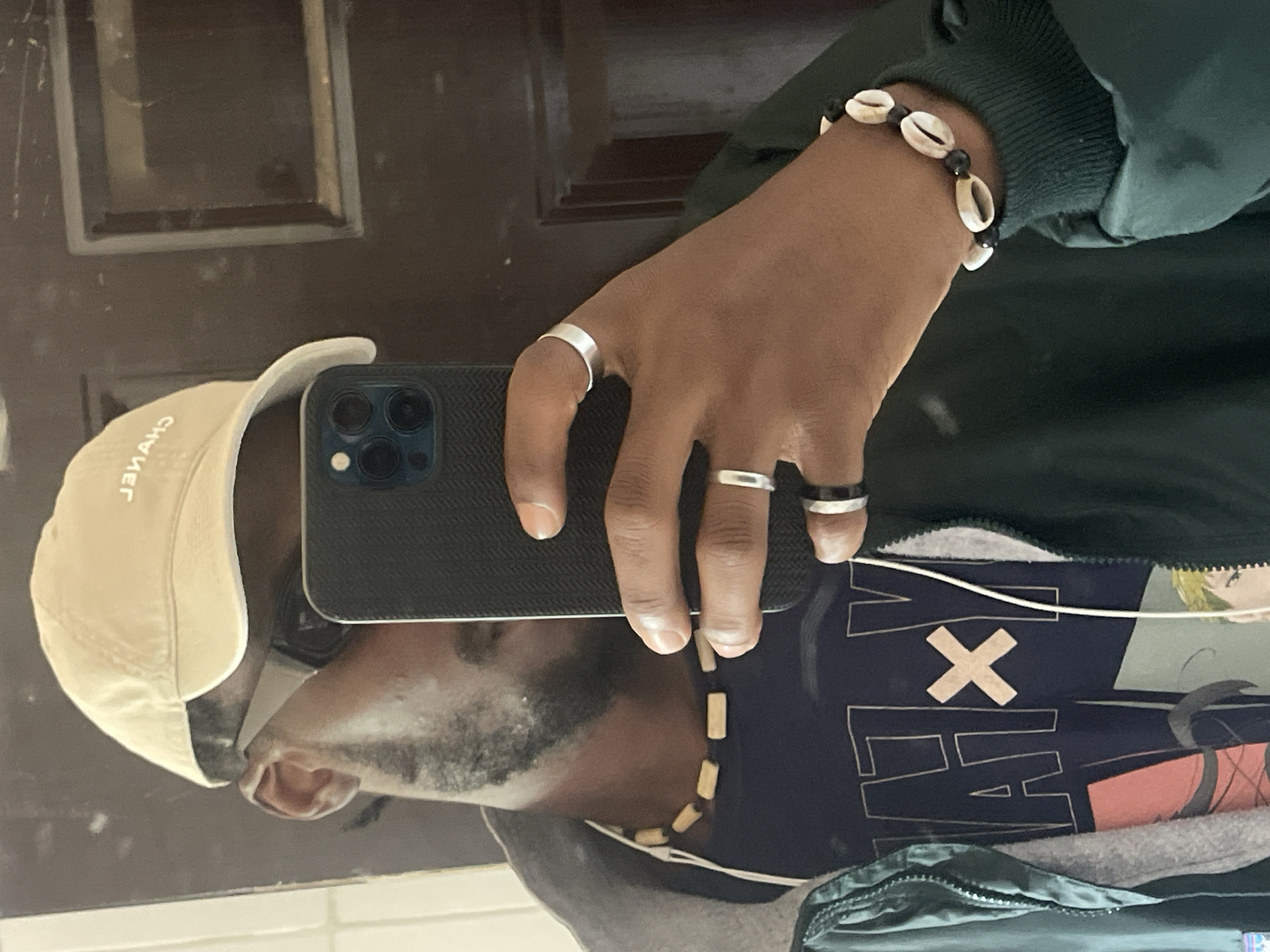 A young Black man with a beard is reflected in a mirror, taking a selfie with his phone. He is wearing a light-colored baseball cap with "CHANEL" written on it, dark sunglasses, and a dark jacket over a navy blue t-shirt. Around his neck is a wooden bead necklace. He has several rings on his fingers and a bracelet with cowrie shells and black beads on his wrist. The background appears to be an indoor setting with a dark wooden door. The image does not contain any identifiable features that would suggest a specific location within the city of Jos, Nigeria.