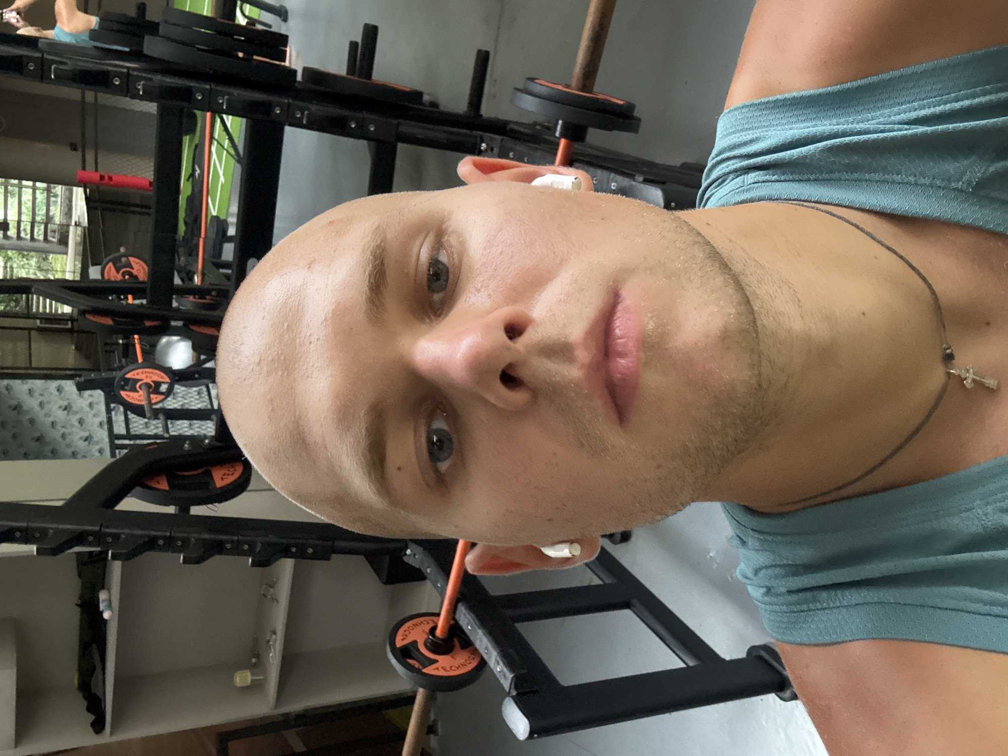 A man with a shaved head and a teal tank top is taking a selfie in a gym. He is wearing white wireless earbuds and a thin necklace with a cross pendant. In the background, there is gym equipment, including a squat rack with weight plates. The environment appears to be an indoor gym.