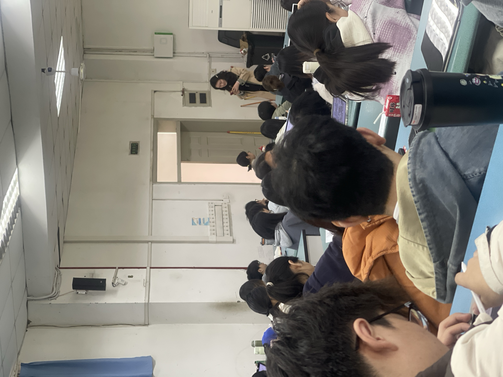 The image depicts a classroom scene in what appears to be Chongqing High-tech Zone, China. A group of students is seated at desks, facing the front of the room. At the front, an individual is standing, possibly teaching or presenting. The students appear to be engaged in a lecture or presentation. The classroom has white walls, with visible fixtures and monitors. There are rows of desks, suggesting it is a lecture hall or classroom. The overall scene suggests a typical educational setting.