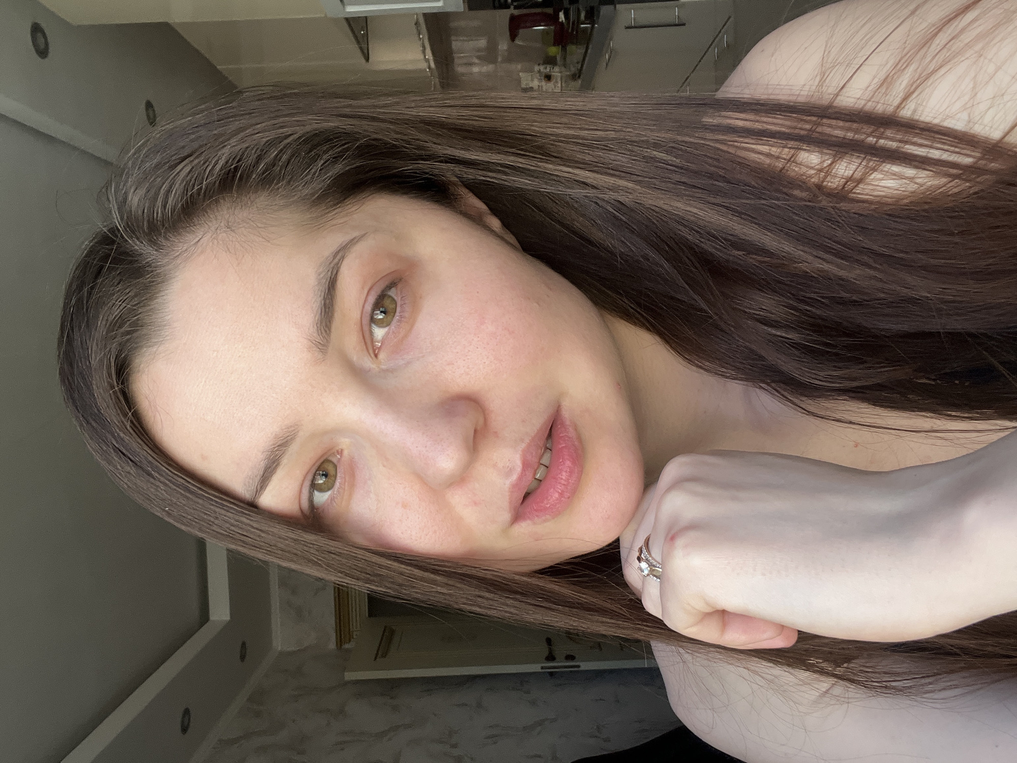 A close-up portrait depicts a person, likely a woman, with long, dark brown hair and hazel eyes, looking directly at the camera. The image is oriented sideways, with her head tilted, appearing to rest on her right hand, which is partially covered by her hair.
Her complexion is light, displaying natural skin texture, including visible pores and some minor redness or freckles on her cheeks and chin. Her eyebrows are dark and well-defined, and her lips are naturally pink and slightly parted, revealing a glimpse of her teeth. She appears to be wearing minimal or no makeup.
On her right ring finger, she wears a silver-toned ring adorned with a central light-colored stone and smaller stones around it. Her long hair cascades around her head and arm.
The background is an interior space, blurred, but shows hints of a modern home setting with neutral-colored walls (grey) and a ceiling featuring recessed lighting. The lighting appears soft and diffused, suggesting natural daylight indoors. No specific activity is taking place other than posing for the photograph. The expression is calm and neutral. There is no visible text. The location context is Khimki, Russia.