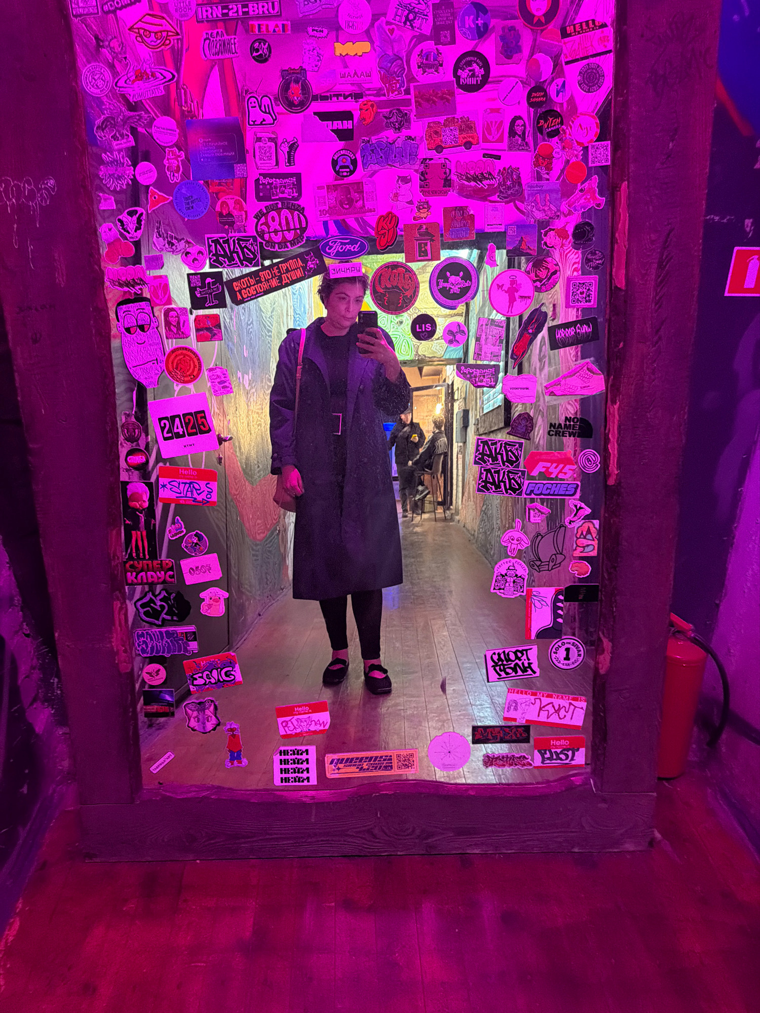 The image depicts a person standing in a narrow hallway, likely in a bar or club, illuminated by purple and pink neon lights. The walls are heavily adorned with stickers of various designs, including cartoon characters, text, and abstract patterns. The person, a woman with short dark hair, is wearing a dark trench coat, black leggings, and black shoes with red accents. She is holding a smartphone and taking a selfie in a large mirror that covers a significant portion of the wall. In the background, other individuals can be seen walking down the hallway. The floor is made of wooden planks. A fire extinguisher is visible on the right side of the image. The overall ambiance is dark and slightly gritty, with a strong emphasis on street art and sticker culture. Given the sticker that reads "Samara" prominently displayed on the wall, this scene is likely located in the city of Samara, Russia.