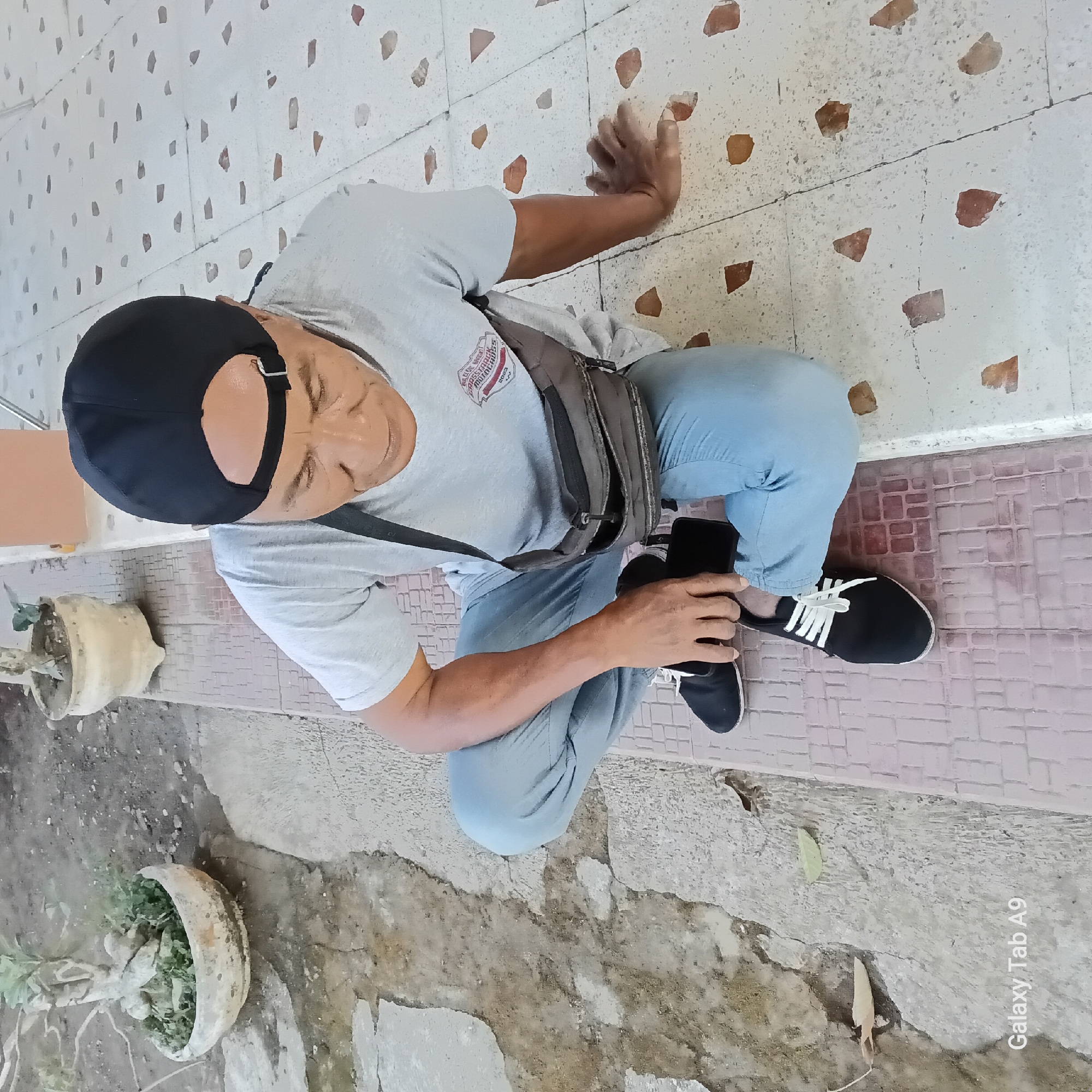 The image shows an elderly East Asian man sitting on a tiled porch in Sambirejo, Indonesia. He is wearing a gray t-shirt, blue jeans, a black baseball cap, and black and white shoes. A dark-colored bag is slung across his chest. The man is holding a black smartphone in his right hand and resting his left arm on the floor. The porch floor is made of white tiles with scattered brown and red chips. To the left of the porch, there is a rough concrete ground with some cracks and dry leaves. In the background, there is a potted plant with a bonsai-like tree. The overall scene appears to be outdoors, in a residential or public area.
