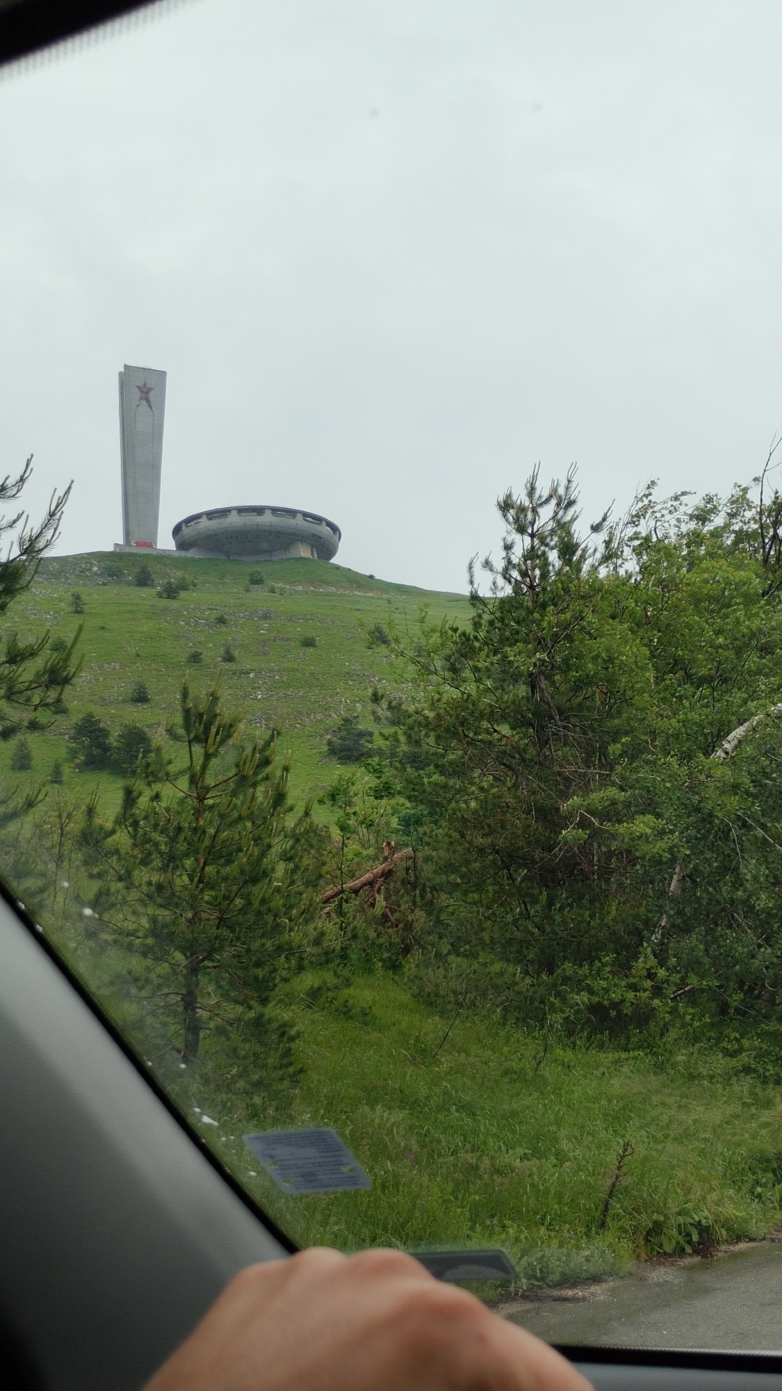 The image depicts a view from inside a vehicle, looking out at the Buzludzha Monument, a historic monument located in central Bulgaria, on the Buzludzha peak in the Balkan Mountains. The monument, with its distinctive saucer-like main structure and tall tower topped with a red star, stands prominently on a green, grassy hill. The sky is overcast and pale. In the foreground, on the right side of the image, is a hand resting on the steering wheel, indicating the perspective of someone driving. A portion of the car's interior, including the dashboard and windshield frame, is visible. Lush green trees and vegetation frame the view of the monument. The overall scene suggests a somewhat bleak, possibly foggy or rainy day.