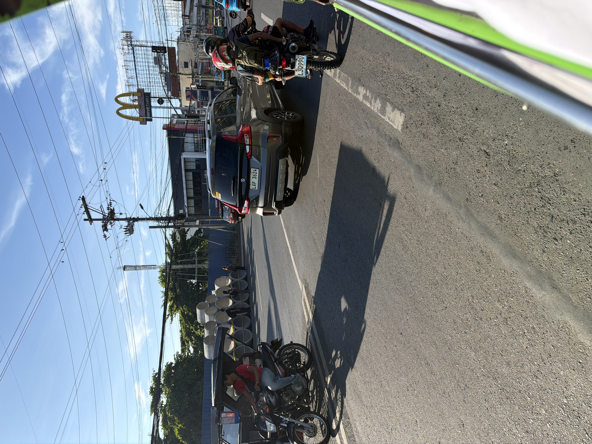 This image captures a street scene in Davao City, Philippines, under a clear blue sky with scattered clouds. The road is paved and appears to be a multi-lane thoroughfare. In the foreground, a motorcycle with a sidecar is stopped, and the driver, a man wearing a red shirt and blue jeans, is seated. Further ahead, a grey SUV with the license plate "LAP 3624" is in motion. Behind the SUV, another motorcycle carrying two individuals, one wearing a red helmet, is also moving.
On the right side of the road, a building with a "McDonald's Drive-Thru" sign is visible. Several utility poles with a complex network of wires are present throughout the scene. To the left, a construction area is indicated by a blue fence and a stack of large concrete pipes. Green trees line the background, interspersed with buildings and more overhead wires. The overall impression is a typical urban environment in the Philippines, with ongoing traffic and commercial establishments.