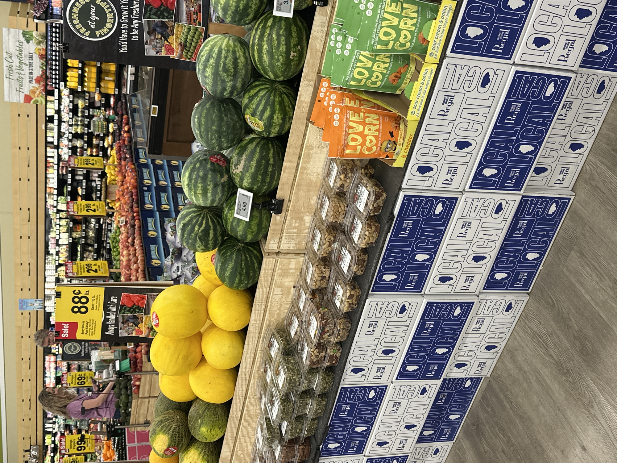 This image depicts the produce and snack aisle of a grocery store, likely a Jewel-Osco, located in Schaumburg, Illinois, United States of America, as indicated by the "Regal Gourmet Snacks" boxes featuring the outline of Illinois. In the foreground, a display of various fruits, including watermelons and cantaloupes, is prominently featured. To the right of the fruits, several boxes of "Love Corn" snacks are visible, with flavors like "Cheddar" and "Sour Cream & Onion." Beneath the fruits, an assortment of pre-packaged nuts and seeds are arranged in clear plastic containers. Behind the produce, shelves are stocked with various items, and several price signs are displayed, indicating sales on items like "Orange Peppers" and what appears to be "Every Bite is Pure Kandy." Two women are visible in the background; one, with curly blonde hair, is interacting with a scanner near a produce display, while the other, with short blonde hair, stands behind her. The overall environment suggests a typical supermarket setting with bright overhead lighting and clean aisles.