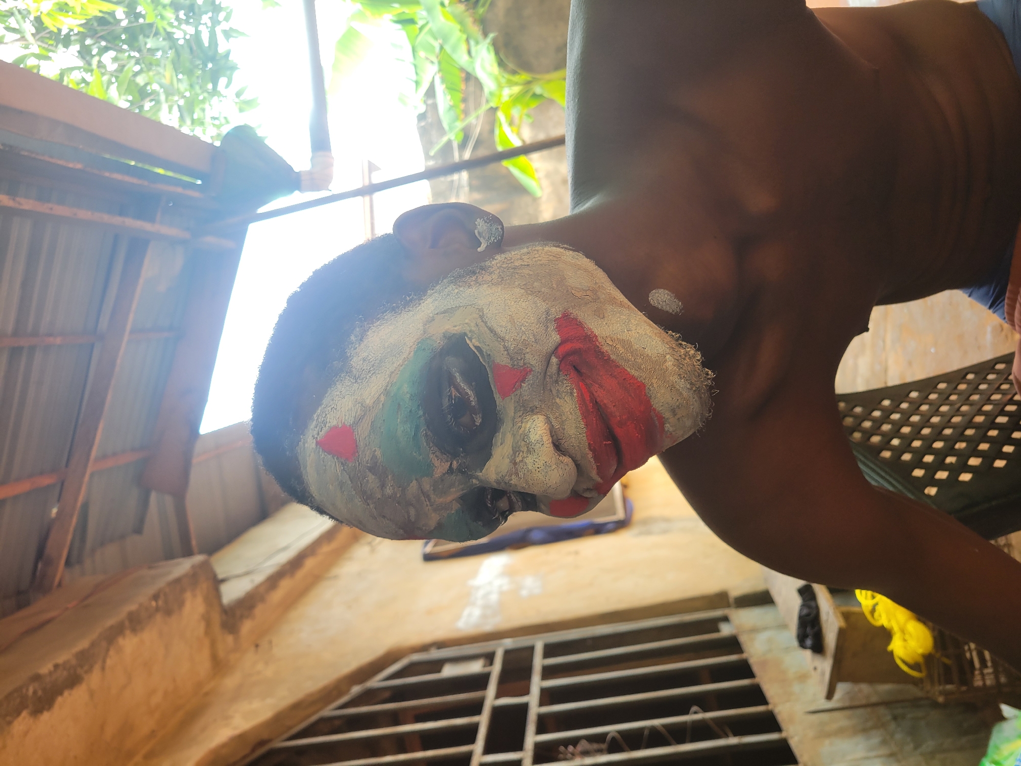 The image shows a person with white face paint, red and green accents, and dark hair, likely wearing a mask or costume. Their torso is visible, with brown skin and a slight sheen, suggesting sweat or oil. The person's head is tilted back, offering an upward angle view of their face. The background includes a corrugated metal roof, some greenery, and parts of a building structure, including what appears to be a ventilation grate and some yellow objects. The setting could be outdoors or in a partially covered outdoor area. No specific location in the city of Unknown, Unknown, can be determined from the image.