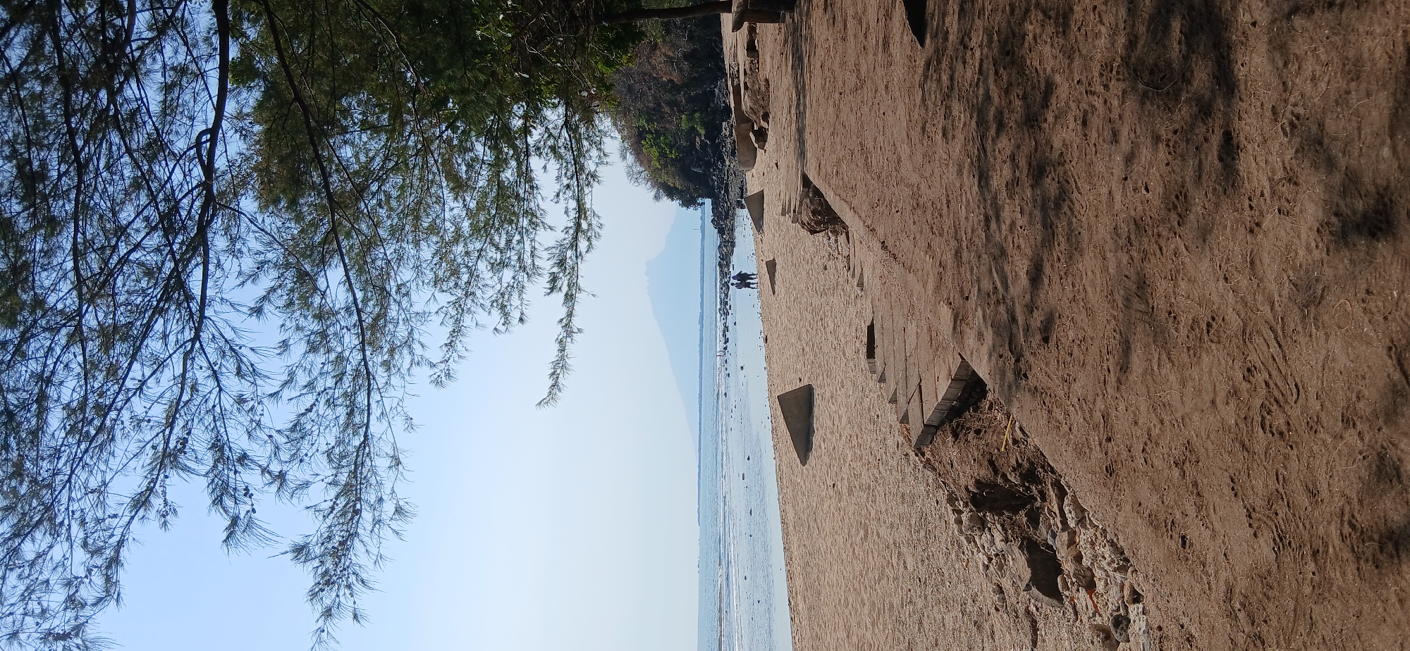 The image depicts a sandy beach in Banyuglugur, Indonesia, under a clear blue sky. In the foreground, the sand is disturbed, showing footprints and some debris. A concrete structure, appearing to be a damaged pathway or steps, is visible along the edge of the beach. The mid-ground features a calm sea with gentle waves lapping the shore. Two figures, a man and a woman, are walking on the wet sand near the water's edge. In the distance, a hazy mountain range forms the horizon, with trees and rocky terrain lining the coast to the right. Lush green trees, with thin, needle-like leaves, overhang the scene from the top of the frame, casting dappled shadows on the sand.