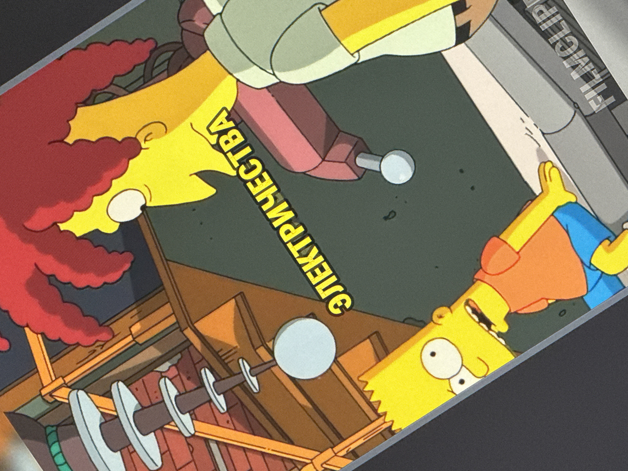 The image depicts a scene from The Simpsons animated television series. Bart Simpson, a young boy with spiky blonde hair, is shown in the foreground wearing an orange t-shirt and blue shorts. He has a surprised expression on his face. Behind him, to the right, is Sideshow Bob, a tall, yellow-skinned character with bright red hair, wearing a light green shirt. A Russian phrase, "ЭЛЕКТРИЧЕСТВА" (which translates to "electricity"), is written in yellow letters across the scene. In the background, there are elements of industrial machinery and brick structures. The image is a photograph of a screen or print, as evidenced by the presence of the watermark "FILMCLIP" in the bottom right corner. There is no information in the image to suggest a location in the city of Saratov, Russia.
