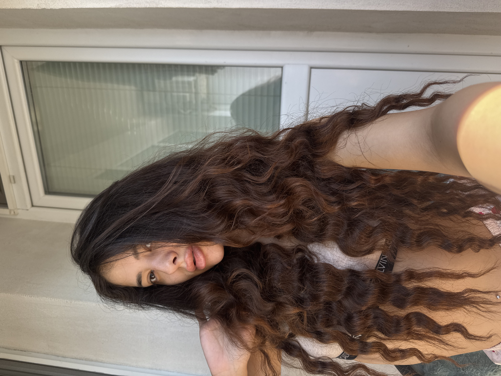 A young woman with long, wavy dark brown hair poses for a photo on a balcony in Plovdiv, Bulgaria. She is wearing a light-colored bra with a black band and what appears to be patterned underwear. Her left arm is raised and her hand is behind her head, her fingers touching her hair. She is looking at the camera with her mouth slightly open. The background shows a white door with blinds and a textured wall. The sunlight casts shadows on the wall and the door.