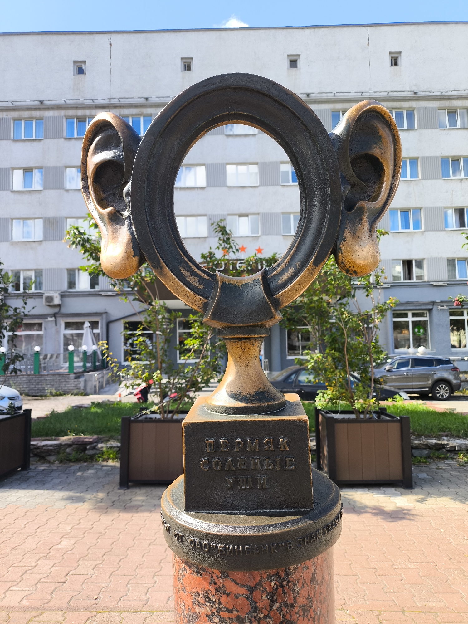 The image depicts a sculpture of a large ear with a ring in the center, located in the city of Perm, Russia. The sculpture is made of metal and is positioned in front of a multi-story building. The building has a light-colored facade with numerous windows and is likely an apartment building or office complex. In front of the building, there are planters with green trees and shrubs. The ground in the foreground is paved with red bricks. There are cars parked on the street behind the sculpture. The sky is blue with some clouds. The sculpture has an inscription in Russian on its base. The overall scene suggests an urban setting in Perm.