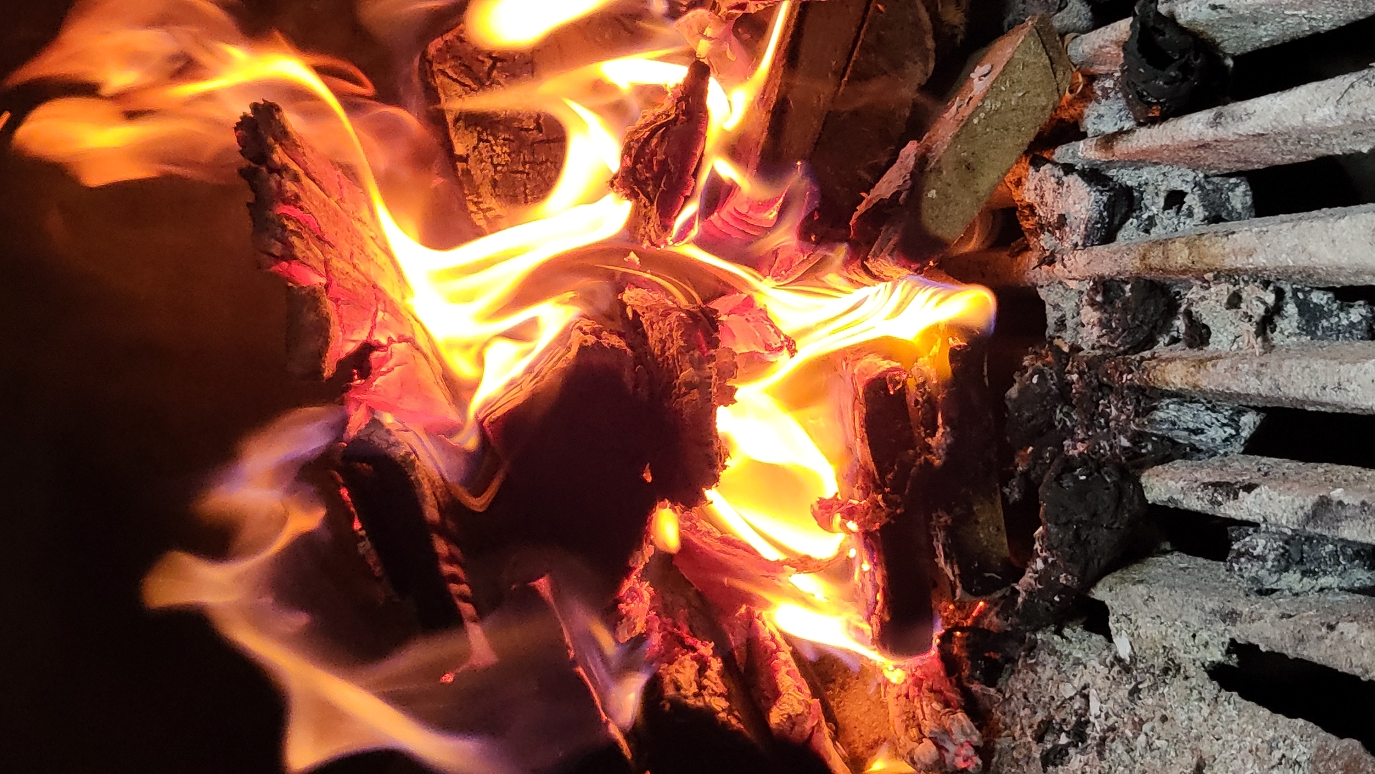 A close-up, vertical, medium shot captures an active campfire with logs burning atop a grill grate. The flames are vibrant orange and yellow, with hints of blue and purple at the edges, licking upwards around the charred wooden logs. The logs themselves show signs of combustion, with glowing red embers and blackened, cracked surfaces. Beneath the burning wood, a thick layer of gray ash and charcoal covers the ribbed metal grill grate, which extends into the foreground. The background is dark and indistinct, suggesting the absence of ambient light. The scene appears to be set outdoors, likely in a backyard or designated fire pit area, and it is not possible to determine the specific location within Prikamskii, Russia based solely on the visual information presented. There are no people or interactions depicted in the image.