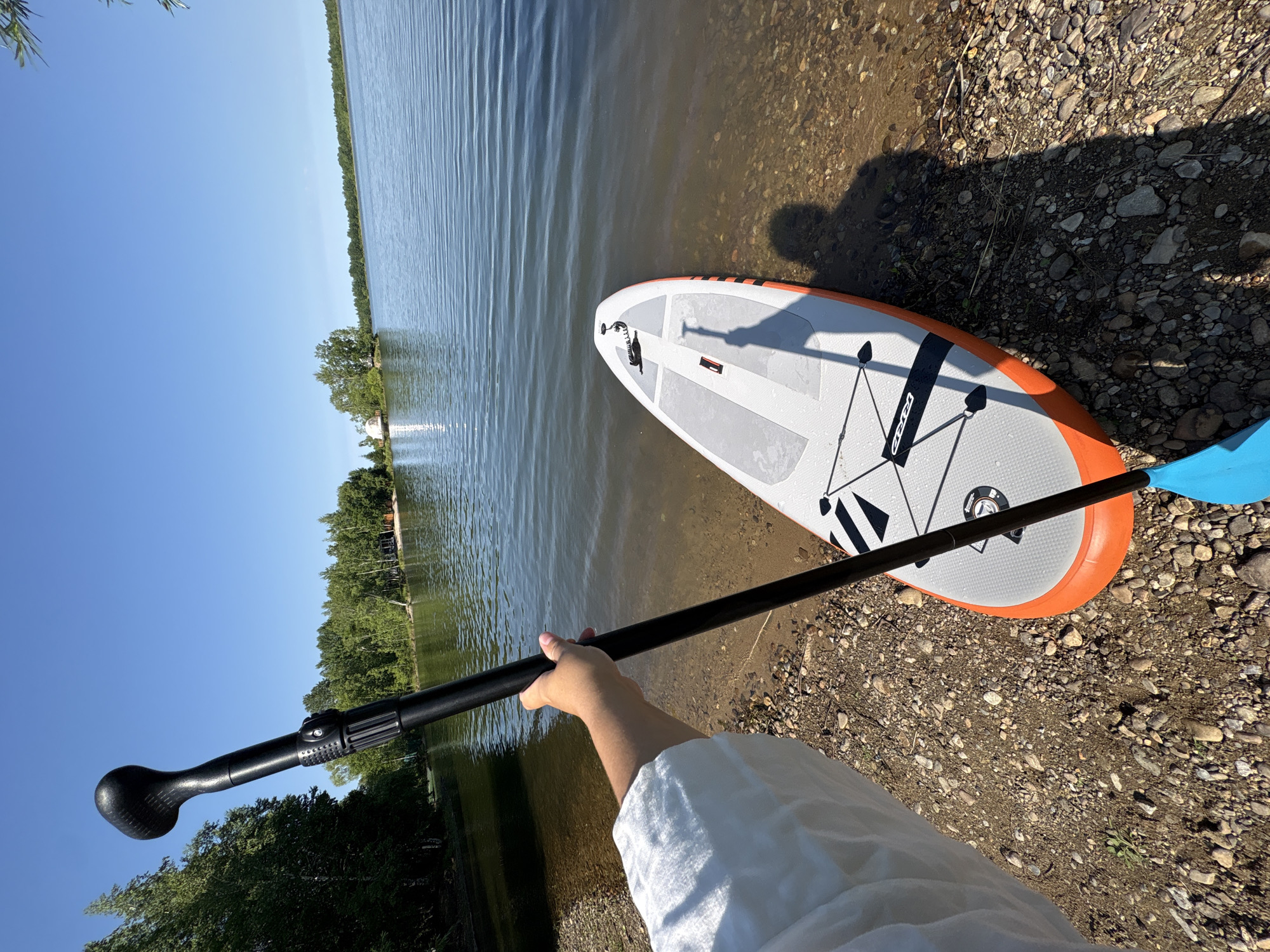 The image depicts a person holding a paddle, standing on the pebbled shore of a calm lake. An orange and grey stand-up paddleboard rests partially in the water, its front end facing the shore. Trees line the far bank of the lake, with a small building visible amongst them. The sky is clear and blue. The location is the city of Razdolnoye, Russia.