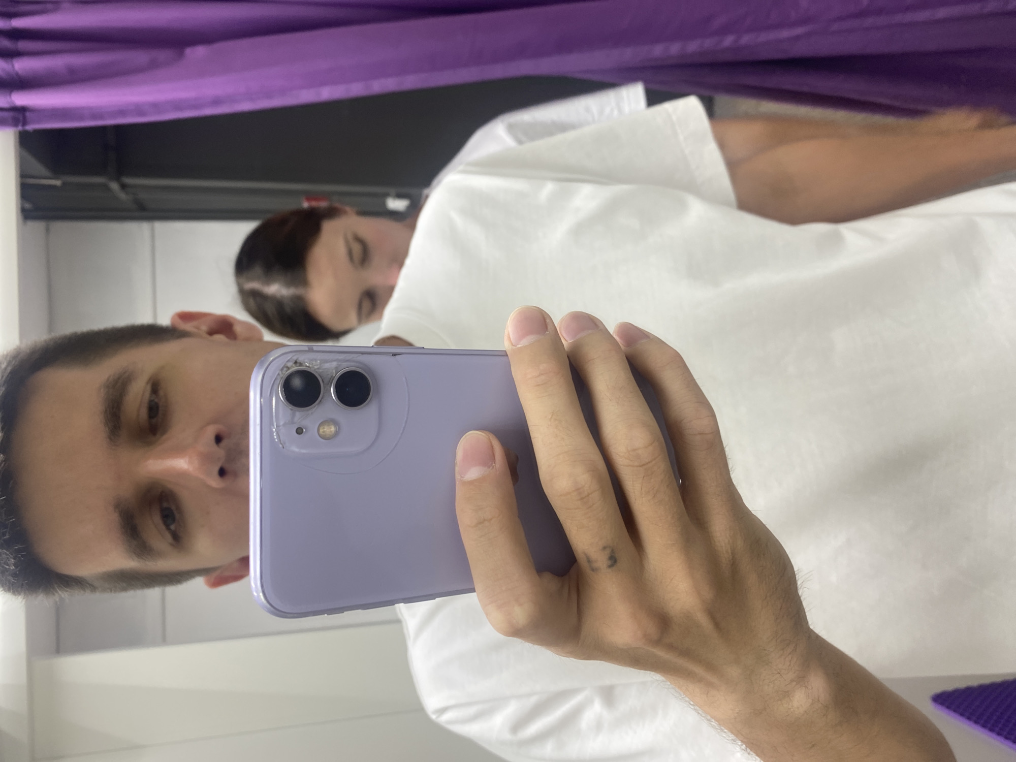 In Novokuybyshevsk, Russia, a person is taking a mirror selfie in a fitting room, holding a lavender iPhone 11. The person is wearing a white t-shirt and has a small tattoo on their finger. In the reflection, another person is visible in the background, also wearing a white t-shirt. The fitting room has a dark background and a purple curtain on the right side.