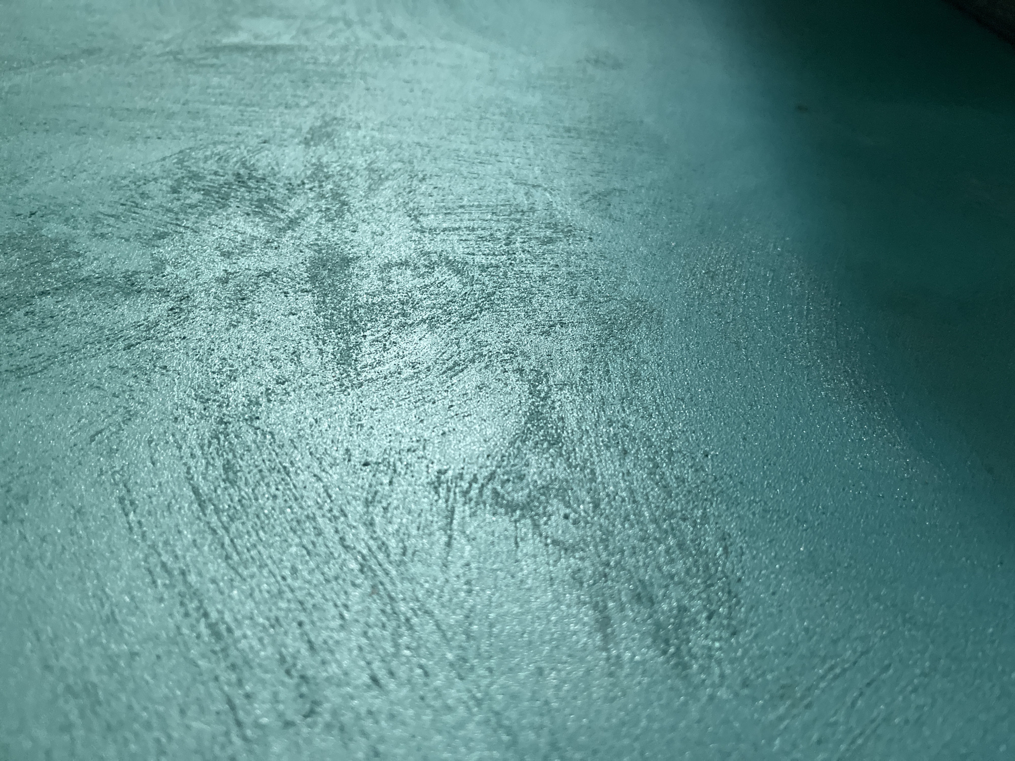 The image presents a close-up, low-angle view of a textured surface, possibly a wall or floor. The dominant color is a muted teal or greenish-blue. The surface exhibits a rough, uneven texture, characterized by granular elements and irregular brush-like strokes that appear to be part of the material or finish. These textural details are more pronounced and in sharper focus in the foreground and towards the left, gradually becoming softer and less defined further into the background and towards the right.
Lighting seems to originate from the upper left, highlighting the raised portions of the texture and creating subtle contrasts across the surface. A darker, less textured area is visible towards the right side of the image, suggesting either a change in lighting conditions or a smoother section of the surface.
No people, specific objects (other than the surface itself), or discernible activities are taking place. The scene is abstract, focusing entirely on the material's surface properties. There are no visible location cues within the image, although the provided context indicates the location is Pokrovnik, Bulgaria. No text is visible. The image conveys a sense of stillness and tactile quality through its detailed depiction of the surface.