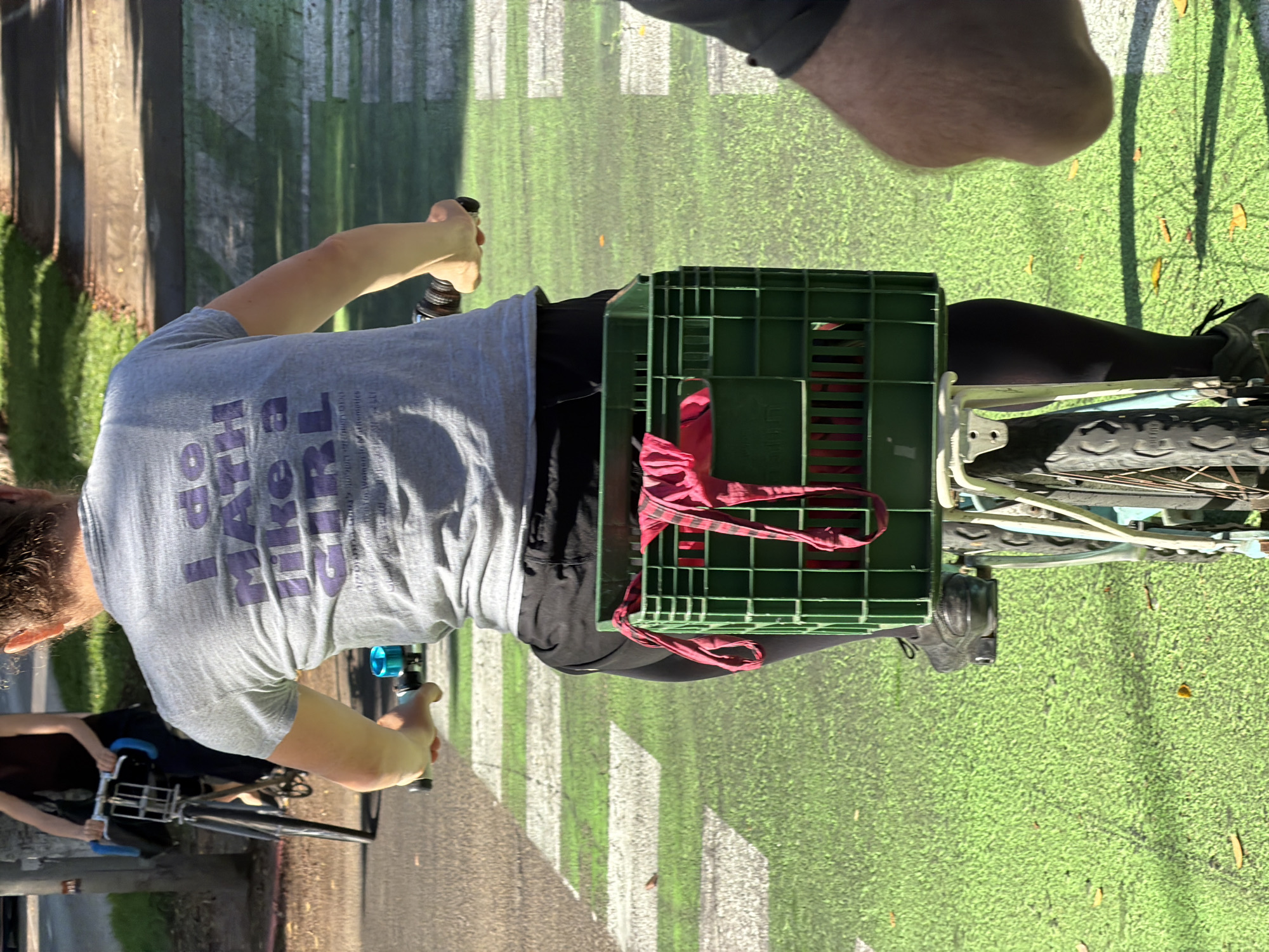 A person rides a bicycle on a green bike lane with white stripes. The person wears a grey t-shirt with purple text that reads "I do math like a girl." They also wear black leggings and black shoes. A green plastic crate is attached to the back of the bicycle, containing a red piece of fabric. Another cyclist is visible in the background, riding a bicycle with blue handlebars. The image appears to be taken on a sunny day, with shadows cast on the ground. The bike lane is part of a city street, and the surrounding environment suggests an urban setting. The distinctive green color of the bike lane is characteristic of several cities in Israel that have invested in cycling infrastructure. Considering the prevalence of such green bike lanes in Tel Aviv, it is highly probable that this image was captured in the city of Tel Aviv, Israel.