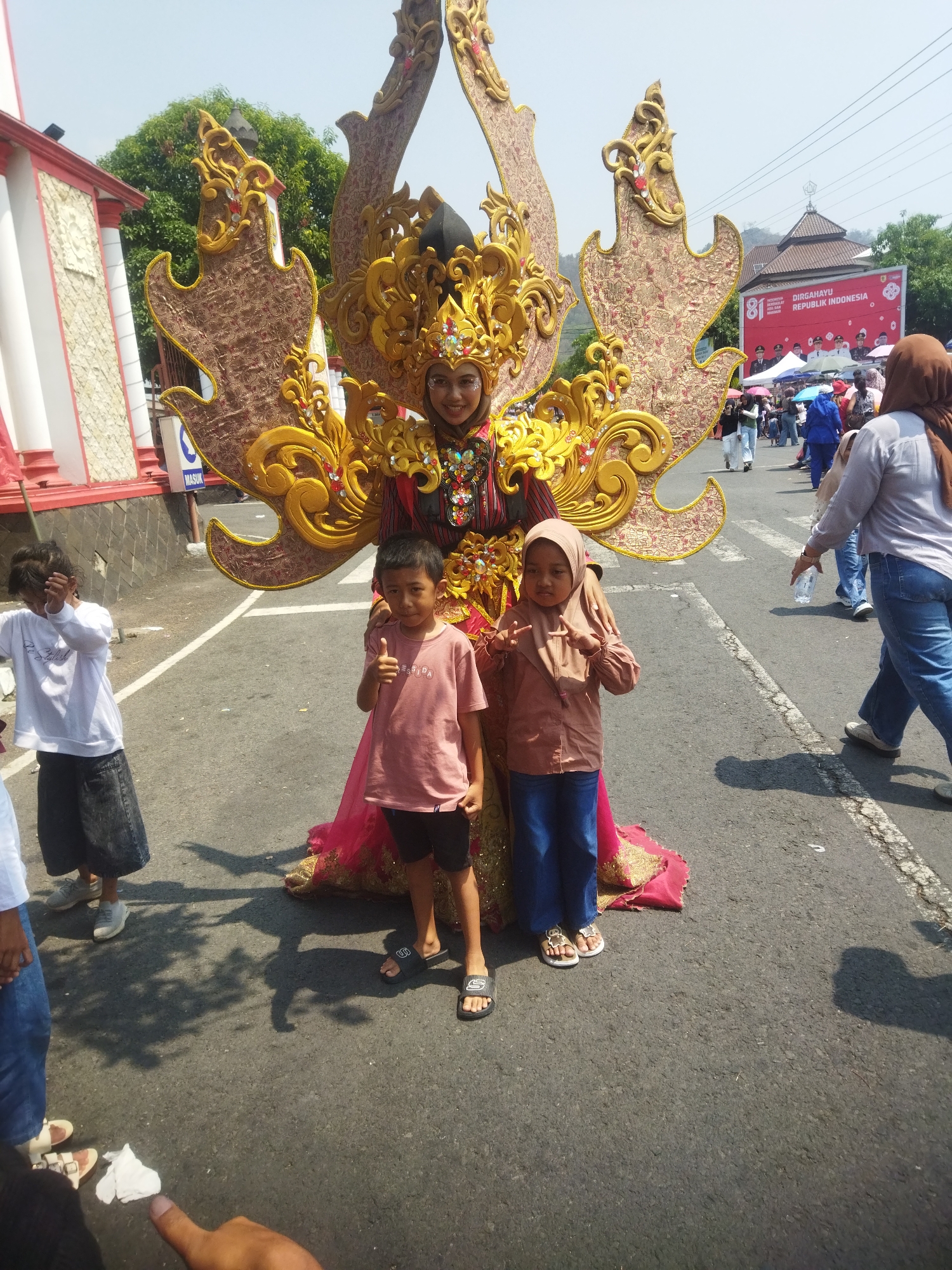 In Wonogiri, Indonesia, a woman in an elaborate golden headdress and costume stands on a street, possibly during a parade. She is adorned with intricate gold detailing, a red and gold patterned dress, and a large, ornate headdress with wing-like extensions. Two children stand in front of her. A boy wearing a pink t-shirt and black shorts gives a thumbs-up. A girl in a brown hijab and matching shirt, wearing blue pants, poses with two fingers raised. Behind them, a crowd watches, and a banner in the background reads "DIRGAHAYU REPUBLIK INDONESIA" with the number "81". The scene appears to be outdoors on a sunny day, with trees and buildings visible in the distance.