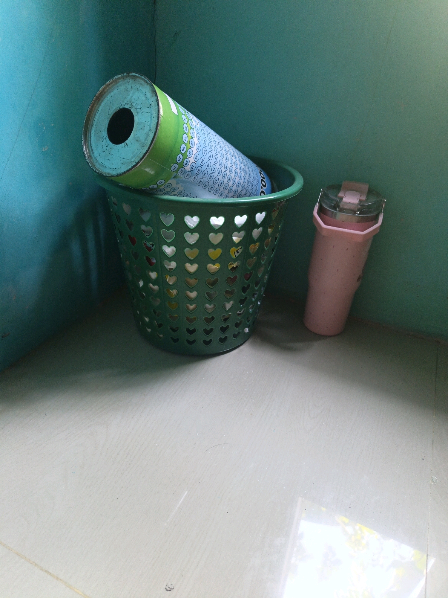 In a corner of a room with teal-colored walls and a tiled floor, a green plastic basket with a heart-shaped cutout pattern rests. Inside the basket is a large cylindrical object, possibly a rolled-up poster or mat, with a green and blue patterned exterior and a visible black opening at one end. To the right of the basket, a tall, pink, octagonal-shaped tumbler with a silver band and a clear plastic lid is leaning against the wall. The lighting suggests an indoor setting, possibly a storage area or a less-used part of a home, without any signs of activity or human presence. The scene is devoid of any specific indicators that would place it in Kamulyan, Indonesia.