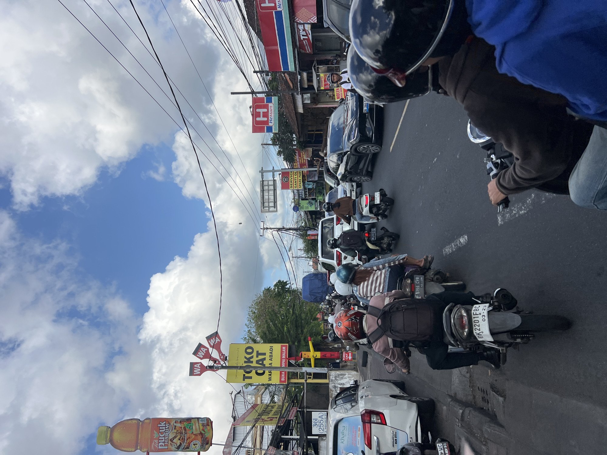 The image shows a street scene in Denpasar, Indonesia, with heavy traffic on a cloudy day. Several motorbikes are visible, some with riders wearing helmets. Cars are also present, including a white SUV and a black sedan. Buildings line the street, with various signs and advertisements displayed, including a large bottle of "Teh Pucuk Harum" and a "HeMart" store. Power lines are strung overhead. The street appears to be busy with commercial activity and transportation.