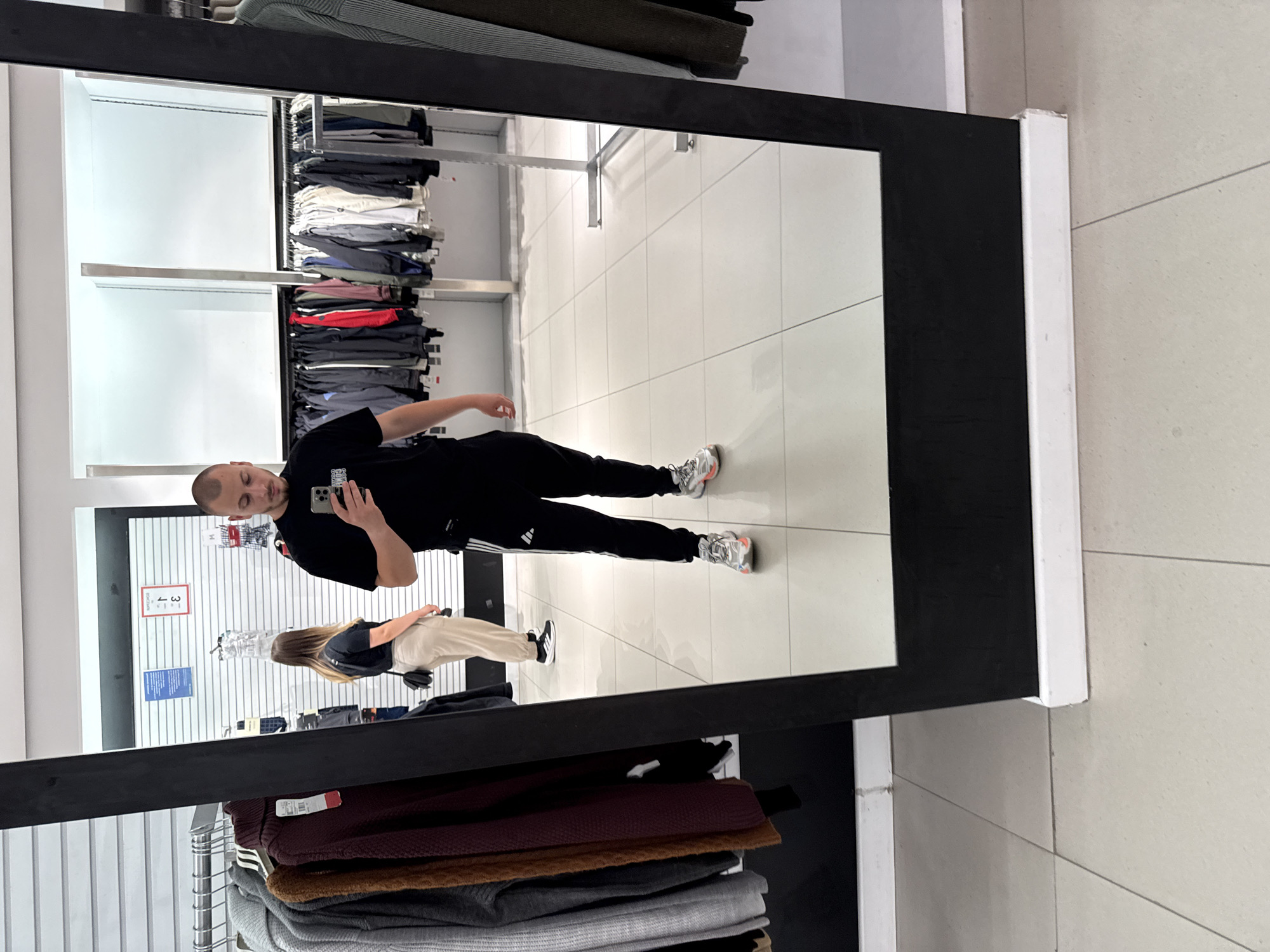 A man is taking a mirror selfie in what appears to be a clothing store in Stara Zagora, Bulgaria. He is wearing a black t-shirt with white stripes on the sleeves and black sweatpants with white stripes. He is also wearing athletic shoes. In the mirror, a woman is visible in the background, wearing a black t-shirt and light-colored pants. She is looking away from the camera. The store has racks of clothing visible, and the floor is tiled. The mirror is large and framed in black. The overall lighting is bright, typical of a retail environment.