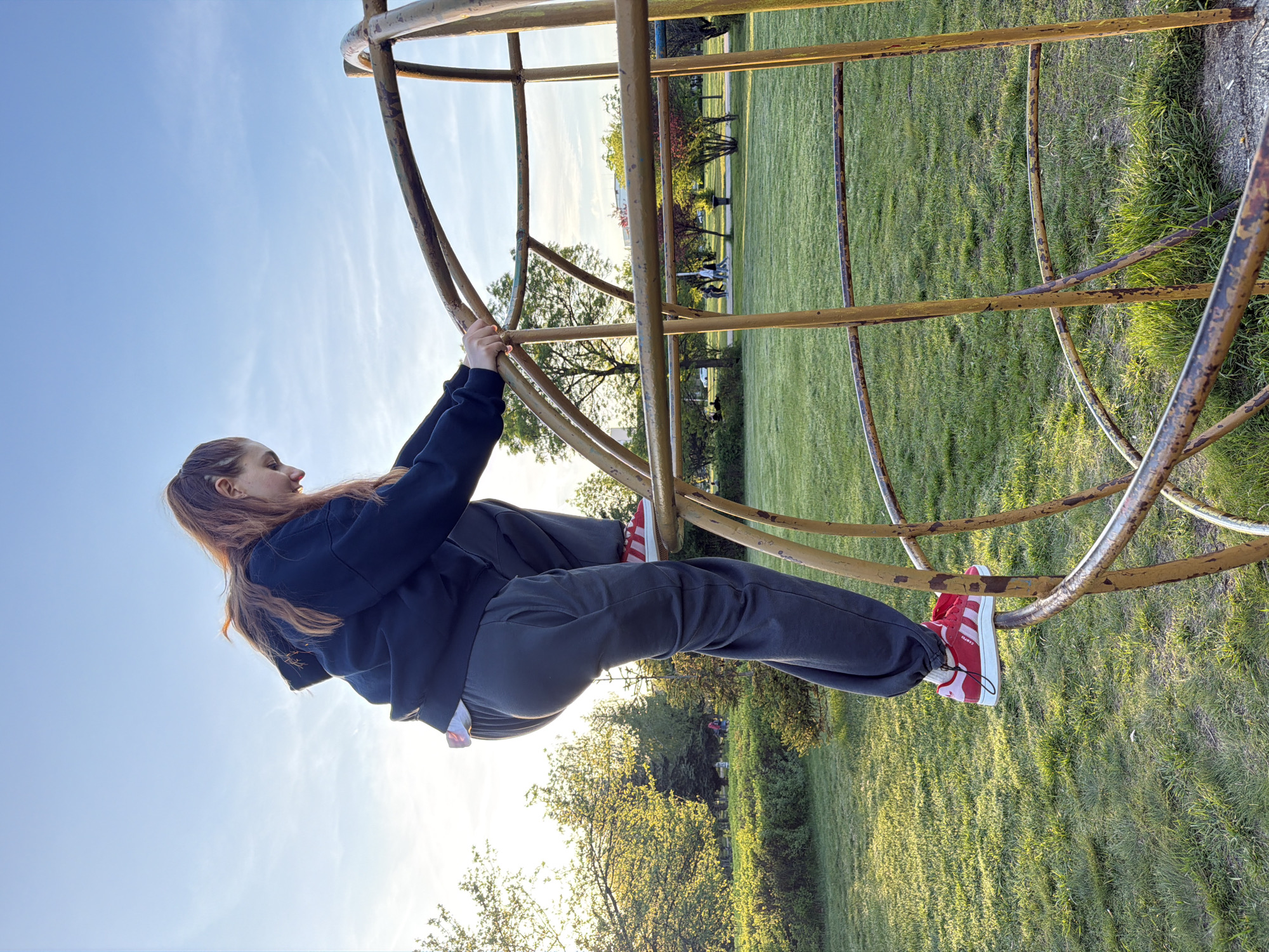 A young woman is depicted actively hanging from a rusty, yellow-painted metal monkey bar structure in an outdoor park. She has long, reddish-brown hair pulled back, and is dressed in a dark blue long-sleeved top and dark grey sweatpants. Her hands firmly grasp two horizontal bars above her, with her body suspended and her feet resting on a lower bar. On her right foot, a distinctive red and white sneaker is visible, with the word "CAMPUS" clearly printed on its side, identifying them as Adidas Campus shoes.
The setting is a large, open park or field, characterized by lush green grass that extends into the background, where several trees, some with new spring foliage, are visible. The sky is a clear, bright blue with faint white clouds. The soft, warm light suggests the time of day is late afternoon or early evening, creating a pleasant and bright atmosphere. The overall scene conveys a sense of recreation and playful physical activity. This park scene is located in Sofia, Bulgaria.
