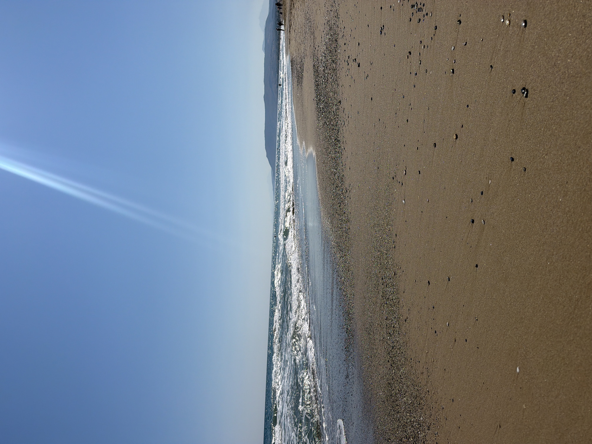 The image depicts a sunny day at a beach in Georgioupoli, Greece. The foreground is dominated by a sandy beach with scattered pebbles and shells. The mid-ground features the Aegean Sea with gentle waves breaking on the shore. The background shows a distant, hilly coastline under a clear blue sky with a prominent sunbeam. Several silhouetted figures can be observed walking and standing along the shoreline in the distance, suggesting a leisurely atmosphere. The overall scene is one of a tranquil coastal environment.