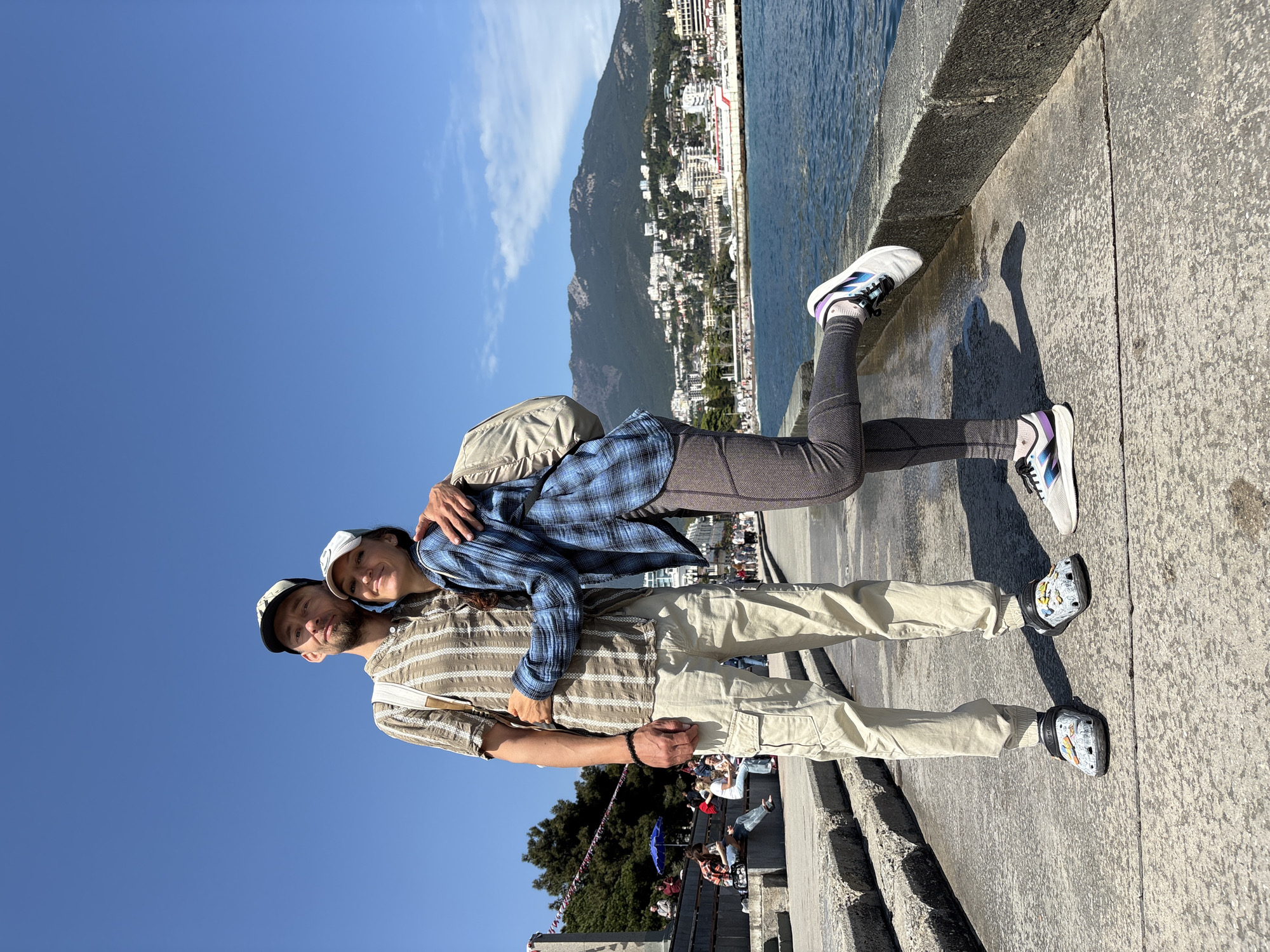 A couple poses in front of a cityscape and water in Ялта, Ukraine. The man wears a striped shirt, tan pants, and Crocs, while the woman wears a blue plaid shirt, leggings, and sneakers. She leans back against him, arm around him. They both wear caps. A mountain with buildings covers the horizon. The couple stands on a concrete surface near the water. In the background, people sit on steps.