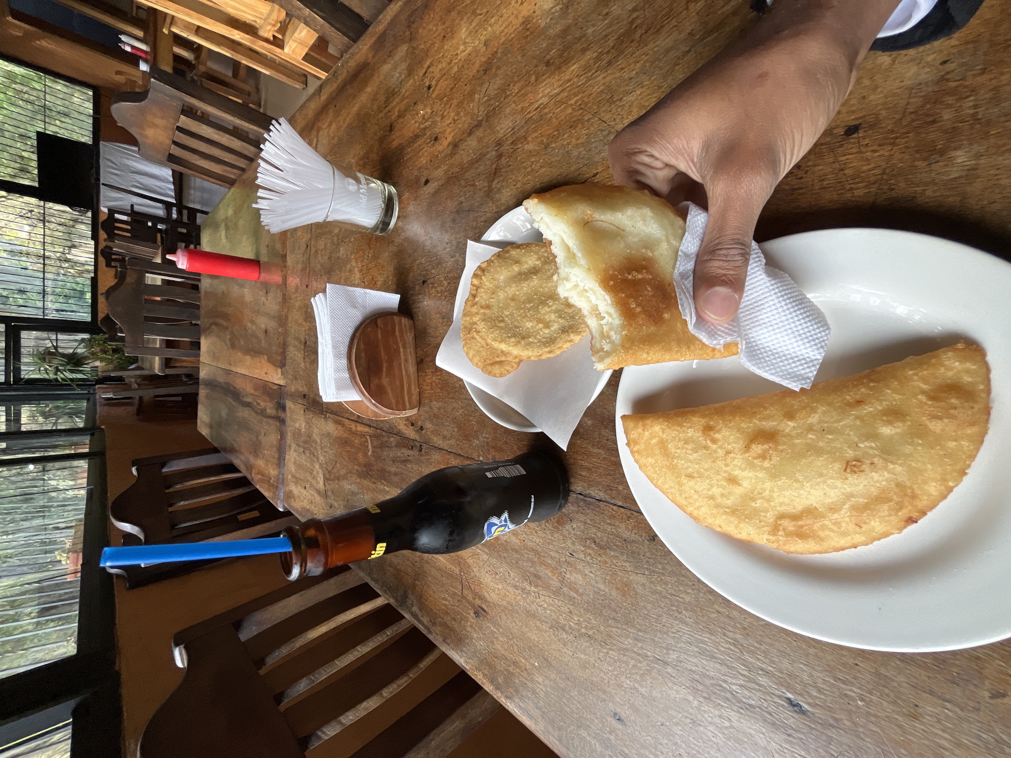 In a rustic restaurant setting, a wooden table displays a meal consisting of two empanadas on a white plate, with a hand holding a partially eaten empanada. Adjacent to the plate are two smaller, round fried pastries, also on a paper liner. A dark bottle with a blue straw stands upright on the table, next to a napkin holder and a glass filled with plastic straws. A red squeeze bottle, likely ketchup, is visible further down the table, with other condiment bottles in the background. The restaurant appears to have large windows overlooking a natural, green environment, consistent with locations in Cacute, Venezuela, a region known for its lush landscapes and agricultural tourism. The lighting suggests an indoor setting with natural light filtering in, and the wooden furniture and rustic decor contribute to a casual, outdoorsy atmosphere.