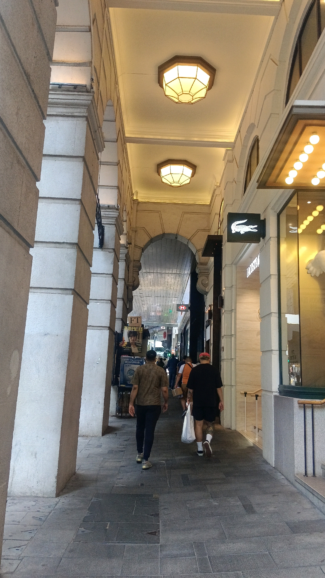 The image captures a pedestrian walkway in Hong Kong, China, characterized by an arched ceiling and classical architectural elements. The walkway is lined with stone columns on the left and shopfronts on the right. Two men are walking away from the camera down the corridor. The man on the left wears a dark brown short-sleeved shirt and dark trousers, and the man on the right wears a black t-shirt, black shorts, and socks with sandals, carrying a white shopping bag. The shops visible include Lacoste, identified by its logo. The ceiling features octagonal light fixtures, and the overall environment suggests a covered shopping arcade or a passageway within a building.