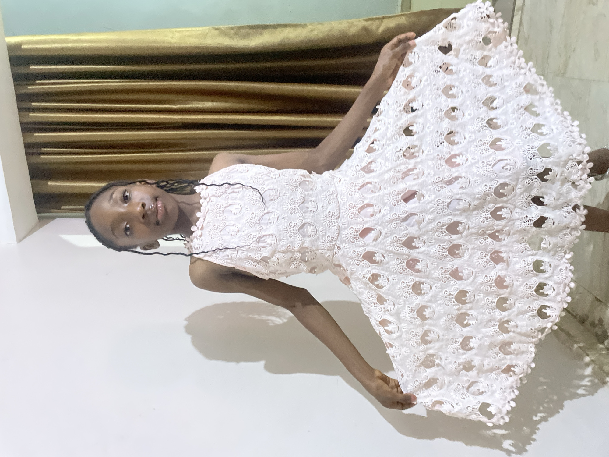 A young African woman is standing in front of a light-colored wall with a gold curtain backdrop. She is wearing a white, lace dress and is holding it out to the sides. The location is likely indoors in Jos, Nigeria.