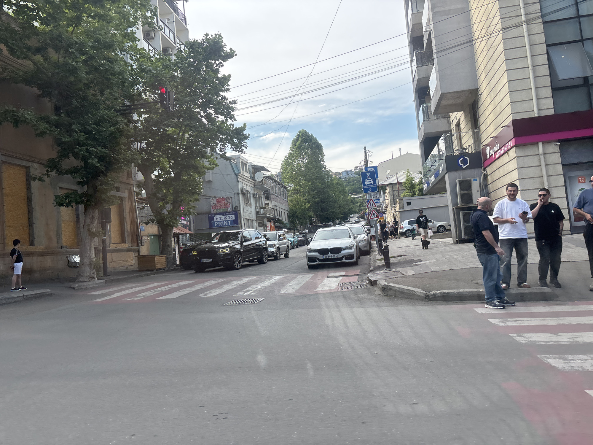 The image depicts a street scene in Tbilisi, Georgia, likely during the daytime. Several vehicles, including a black SUV and a white sedan, are visible on the road, with some stopped at a pedestrian crossing. A traffic light is red, indicating a stop. Pedestrians are present on the sidewalk and crossing the street.
In the foreground, three men stand at a crosswalk, facing away from the camera. Two of them are wearing dark shirts and jeans, while one is in a white t-shirt and dark pants. Further down the street, a young person in shorts and a t-shirt stands on the sidewalk. Buildings line the street, some with balconies and windows. Trees with green foliage are interspersed along the roadside.
Signs are visible on some buildings, with text in Georgian script, including one that appears to say "PRINT". A blue traffic sign with icons, one depicting a car, is also present. The sky is partly cloudy, suggesting a temperate weather condition. The overall atmosphere appears to be that of a typical urban environment with moderate pedestrian and vehicle activity.