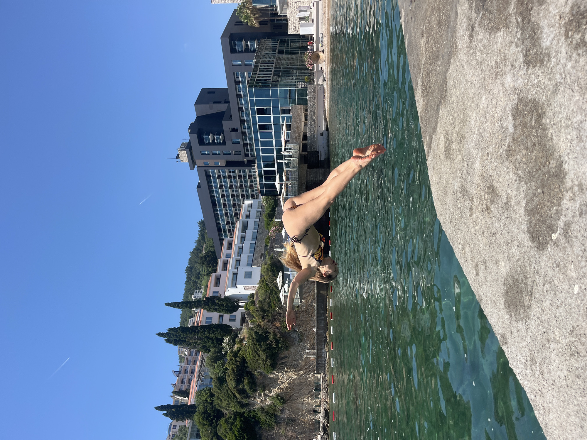 A female diver is captured mid-air, in a graceful dive towards the clear, emerald green waters of the Adriatic Sea. Her body is parallel to the water's surface, with her arms outstretched and legs extended. She is wearing a patterned bikini.
The dive is taking place from a concrete ledge, which occupies the foreground of the image. Behind the diver, a picturesque coastal scene unfolds. The coastline is lined with white buildings, some multi-story, nestled amongst lush green trees and rocky outcrops. In the background, a modern, dark-colored hotel complex with glass-paneled balconies dominates the skyline.
The sky above is a brilliant, cloudless blue, with a faint contrail from an airplane visible in the upper left corner. The water is calm, with gentle ripples reflecting the bright sunlight. Red and white buoys mark a swimming area in the sea. The location is identifiable as the area near the Saint Nicholas Island, a well-known landmark in Budva, Montenegro, a popular tourist destination on the Adriatic coast.