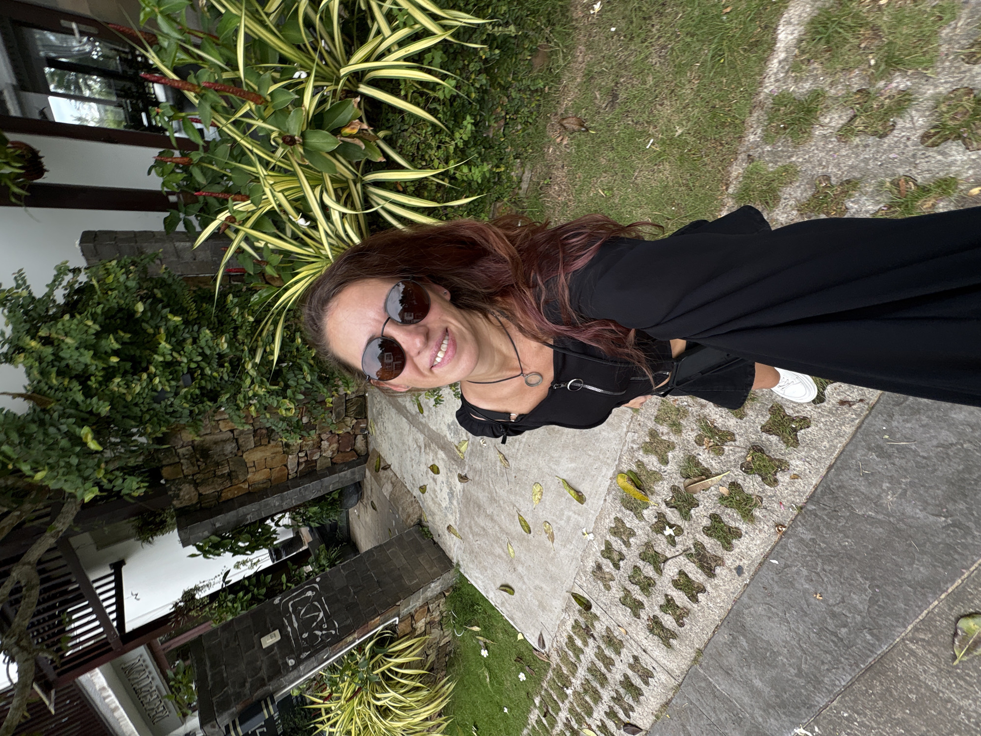 A medium shot captures a woman with long, dark red hair and sunglasses standing outside, posing for a selfie. She wears a black top and skirt, white sneakers, and a silver pendant necklace. Her right hand is out of frame, holding the camera. The background features lush green foliage, a stone wall, and a paved walkway with scattered leaves. To the left, a sign reads "RECEPTION." The environment appears to be a tropical or subtropical location, possibly a resort or hotel.