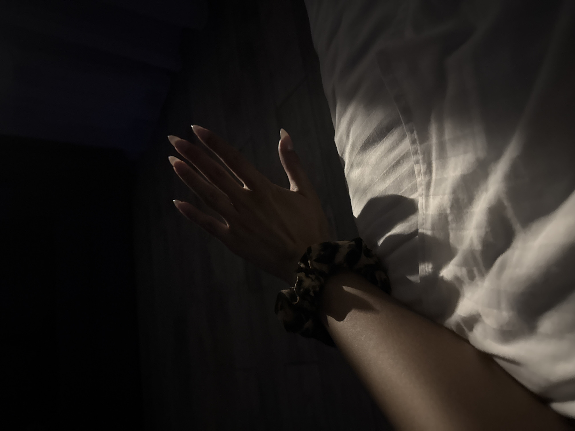 A person's arm and hand are visible in the foreground, resting on white bedding. The arm is adorned with a patterned scrunchie. The hand is open, with fingers extended and painted nails. The scene is dimly lit, with a spotlight illuminating the hand and part of the arm, casting shadows across the bedding. The background is dark and out of focus, suggesting an indoor setting, possibly a bedroom at night. The location in Doma 1096 Km, Russia, indicates a remote or specific address.