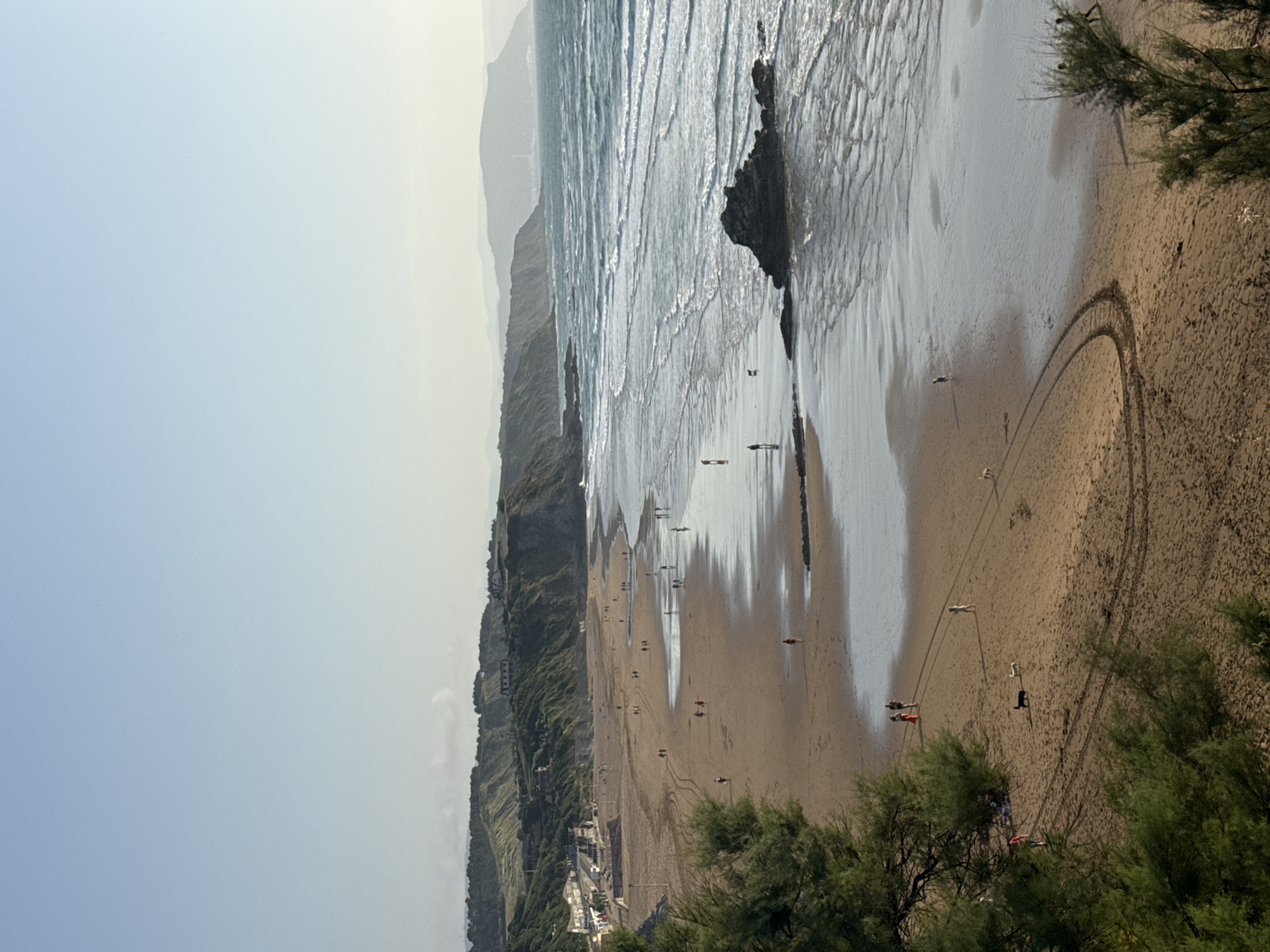 The image depicts a scenic view of the beach in Sopela, Spain, under a clear, pale blue sky. The sandy beach stretches along the coastline, with gentle waves from the turquoise ocean lapping at the shore. Rolling green hills rise behind the beach, dotted with buildings and trees. Several people are visible on the beach, walking, strolling, and some with dogs. A large rock formation is visible in the shallow water. The scene captures a peaceful, late afternoon or early evening atmosphere.