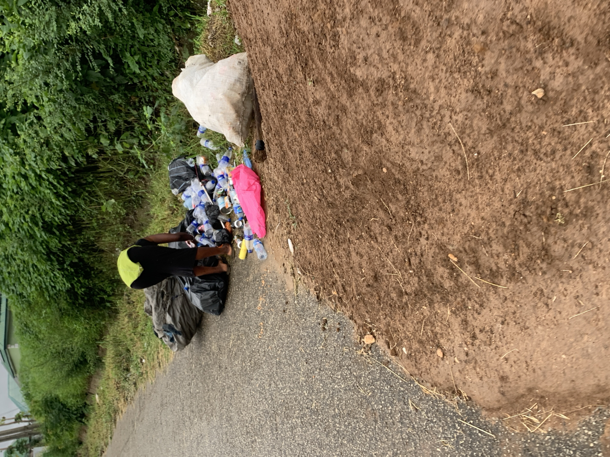 The image captures an outdoor scene in Abuja, Nigeria, featuring a person engaged in collecting or sorting waste by a roadside. The subject, dressed in a dark top with a bright lime-green or yellow cap/hood and dark shorts, is bending over a substantial pile of discarded items. This refuse, situated on the granular, dark grey asphalt of a paved road, includes numerous plastic bottles (predominantly clear and blue, with some yellow), aluminum cans, and multiple black plastic garbage bags. A distinctive bright pink plastic item lies atop the pile.
Adjacent to the waste and the road, a steep, reddish-brown dirt embankment rises, partially covered by sparse dry grass and debris. A large white woven sack, likely used for collecting materials, rests against the base of this embankment. Lush green vegetation, comprising bushes and trees, forms a dense backdrop on the left side of the image, suggesting a natural or undeveloped area. A light-colored building or structure is faintly visible in the far-left background.
The scene takes place during daylight hours, characterized by soft, diffused lighting, indicating either an overcast sky or early morning/late afternoon. No harsh shadows are visible. The activity of the person, surrounded by discarded materials, strongly suggests waste picking or informal recycling. There is no visible text in the image.