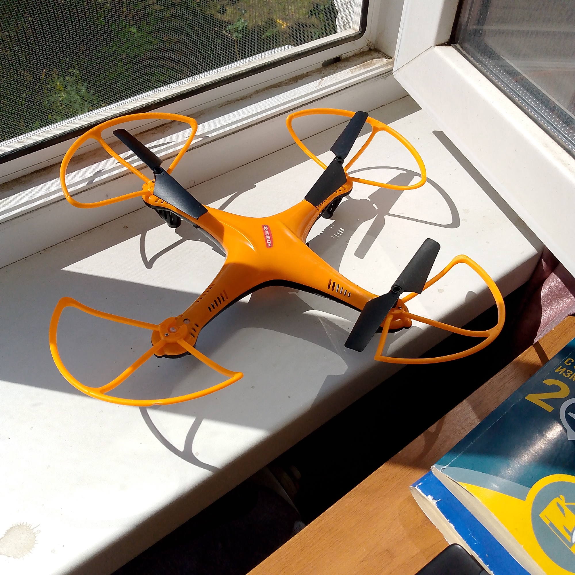 An orange drone with black propellers rests on a white windowsill, which appears to be inside a building. The drone is angled, with its front facing towards the left of the frame. One of its orange propeller guards is prominently visible in the foreground. Sunlight casts shadows from the drone onto the windowsill. Outside the window, a green, leafy area is visible through a screen. To the right of the drone, a stack of books, one with a blue and yellow cover, rests on a wooden surface. The scene suggests a casual placement of the drone, possibly awaiting use or after being taken out. The location of Nikologory, Russia is not ascertainable from the image content.