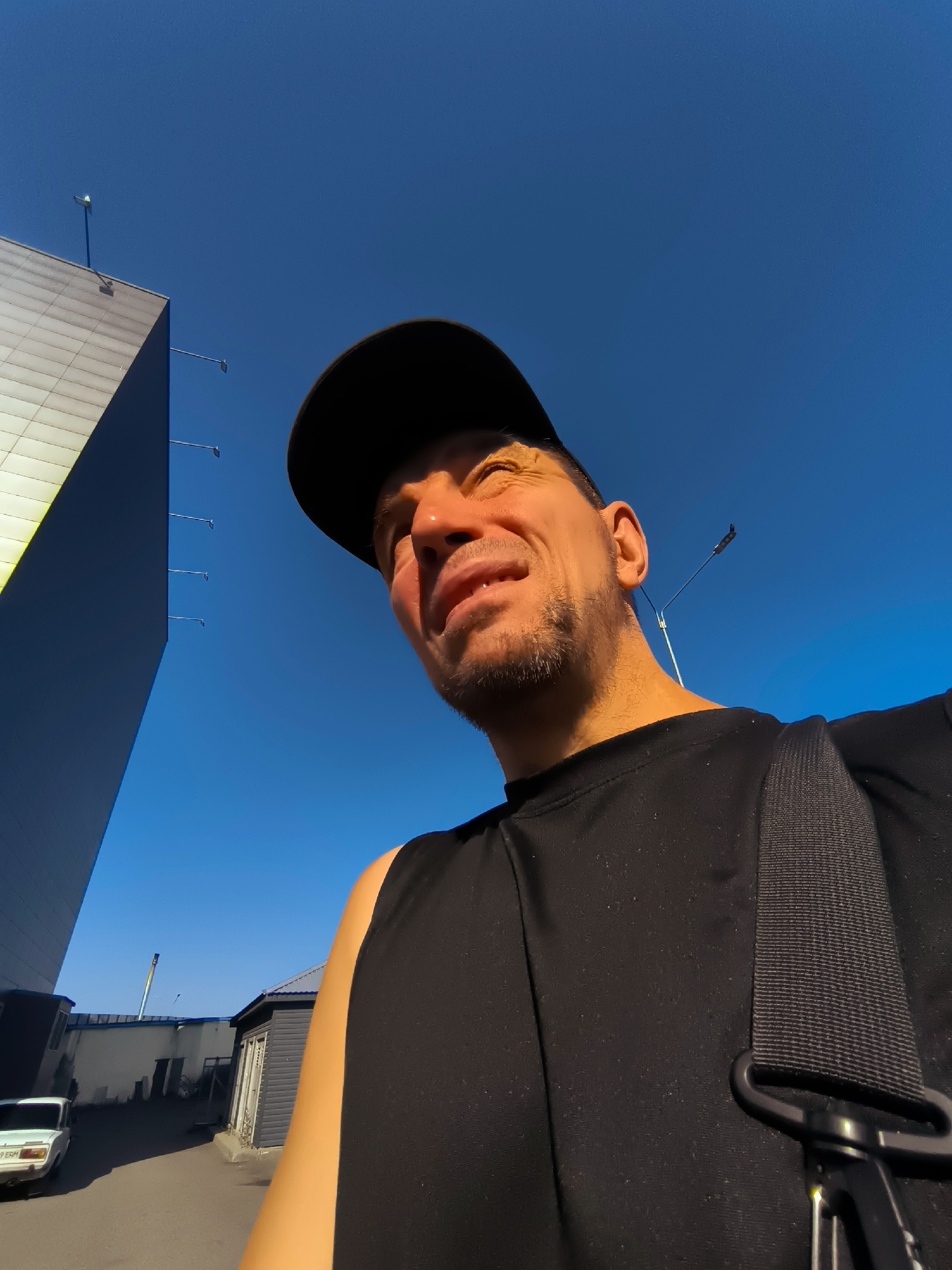 A man wearing a black baseball cap and a black t-shirt is looking upwards with his eyes squinted due to the sunlight. He has stubble on his face. A black strap is visible across his t-shirt, suggesting he is wearing a bag. In the background, a modern, angular building with a pale facade is on the left, with the clear blue sky above. To the right, there are some smaller, dark-colored buildings and a street with a white car parked on the left. The location is identified as being in the city of Qarağandı city administration, Kazakhstan.