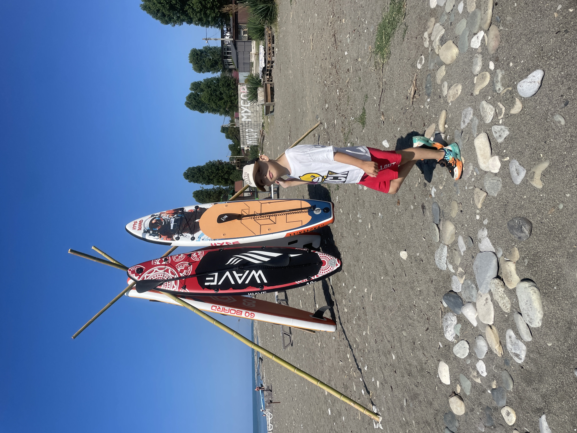 A young boy in a white t-shirt and red shorts stands on a pebble beach next to three paddleboards leaning against bamboo poles. The paddleboards are decorated with various designs, including geometric patterns and an astronaut. The beach is sandy with scattered rocks. In the background, there are trees, buildings, and a clear blue sky. The sea is visible in the distance, with a few people and a boat on the shore. The location appears to be Giachrypsh, Abkhazia, as suggested by the "MYCOP" graffiti visible on a wall in the background, a common sight in the region referring to the local waste management company.