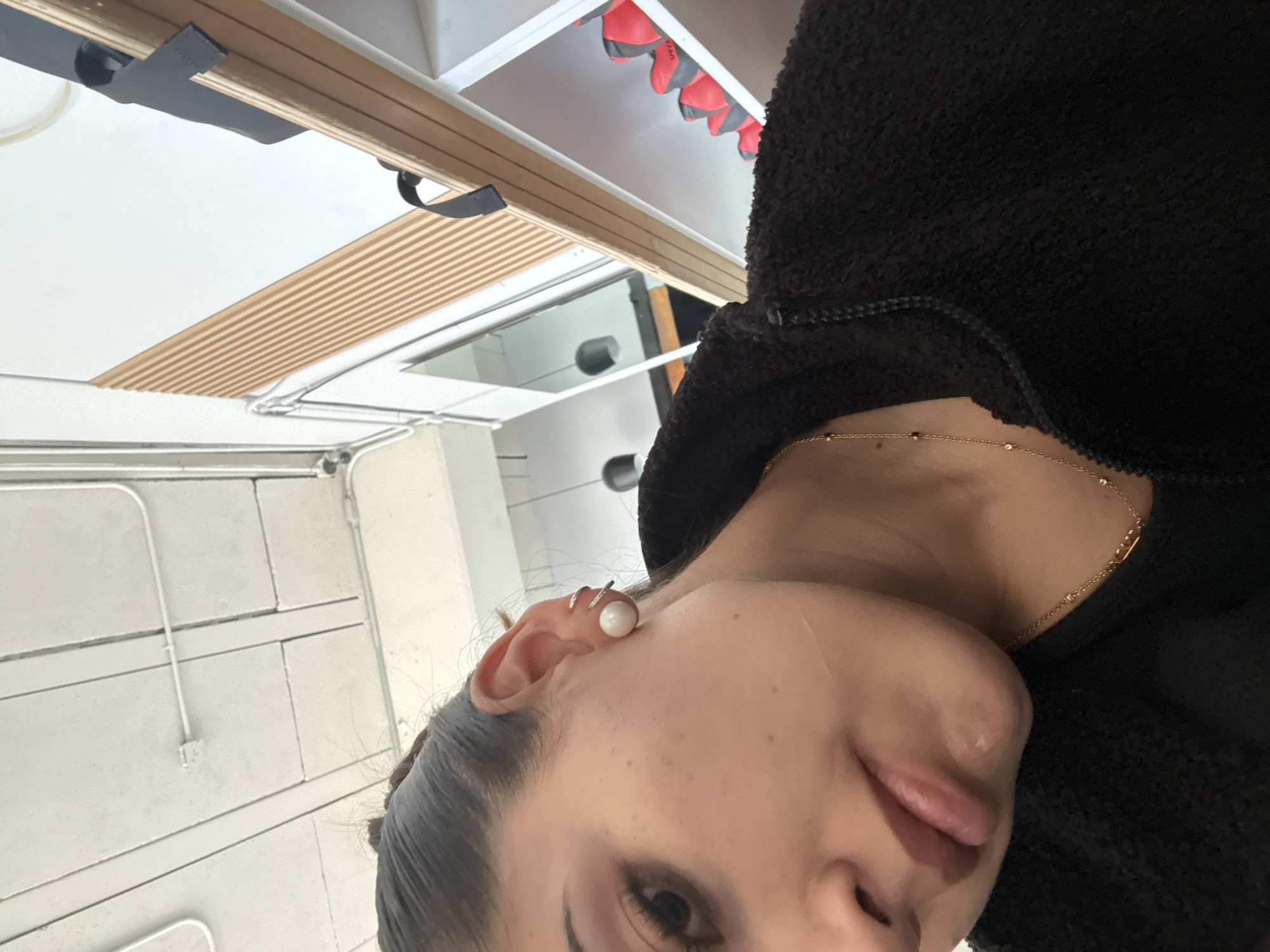 The original image is rotated approximately 90 degrees clockwise, showing a close-up of a person's face from a unique angle. The subject is a young woman, partially visible from her left eye to her neck, with her head tilted. She has dark hair, styled neatly, and wears eye makeup. Her lips are slightly parted, and she is subtly sticking out her tongue, conveying a playful or casual expression.
She is adorned with a prominent round white pearl stud earring in her left ear, complemented by several small silver-colored ear cuffs or hoops higher on her helix. Around her neck, a delicate gold chain necklace with small dark beads or stones is visible. She is wearing a dark, textured garment, possibly a fleece or sherpa-style jacket or hoodie.
The background is an indoor setting with varied architectural elements. To the left of her head (when viewed upright), there is a light-colored wall composed of rectangular panels, traversed by white electrical conduit pipes. To her right and above her, a wooden slatted panel is visible, with a wider wooden beam and black straps above it. Further back and to the right, several red and gray padded objects, possibly gym equipment like punching bags or flotation devices, hang from a ceiling structure. A reflection in a shiny surface, possibly a window or mirror, shows dark, cylindrical shapes.
No discernible text is visible. The industrial yet functional aesthetic of the background elements, coupled with the red and gray objects, suggests a gym, studio, or commercial space, consistent with an urban setting like Puebla, Mexico. The lighting appears to be artificial or diffused natural light.