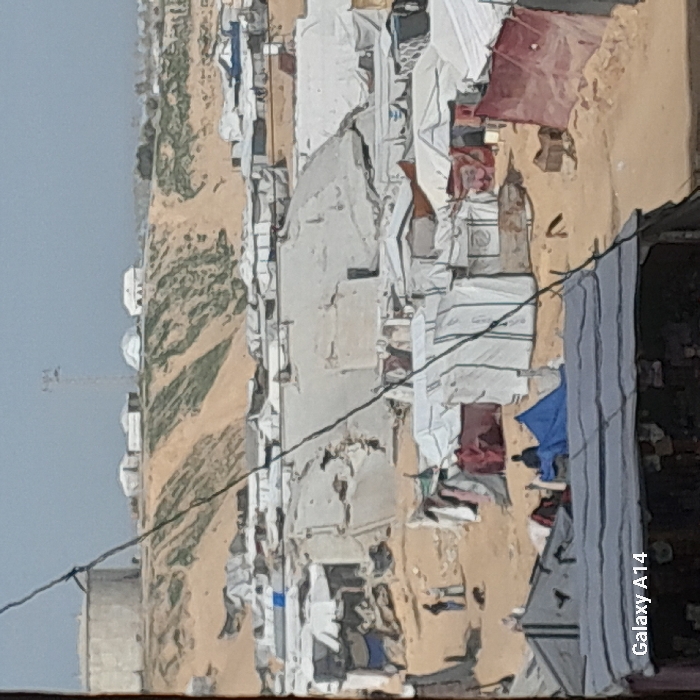 The image displays a densely populated makeshift encampment situated in Khan Yunis, Palestinian Territory, under a hazy sky. A dark cable runs diagonally across the upper-left to lower-right foreground, partially obscuring a dark roof at the bottom. The midground is characterized by numerous temporary shelters and tents, constructed from various materials including white, blue, grey, and red tarpaulins and sheets, spread across sandy, uneven ground. Two individuals stand together in the mid-left area, near a dark animal on the ground, while another person sits on the ground next to a blue-tarped structure slightly further right. The background features an elevated sandy ridge with patches of green vegetation. On this ridge, a large, light-colored cylindrical storage tank is visible on the left, accompanied by several smaller light-colored structures and a tall communications mast to the right.