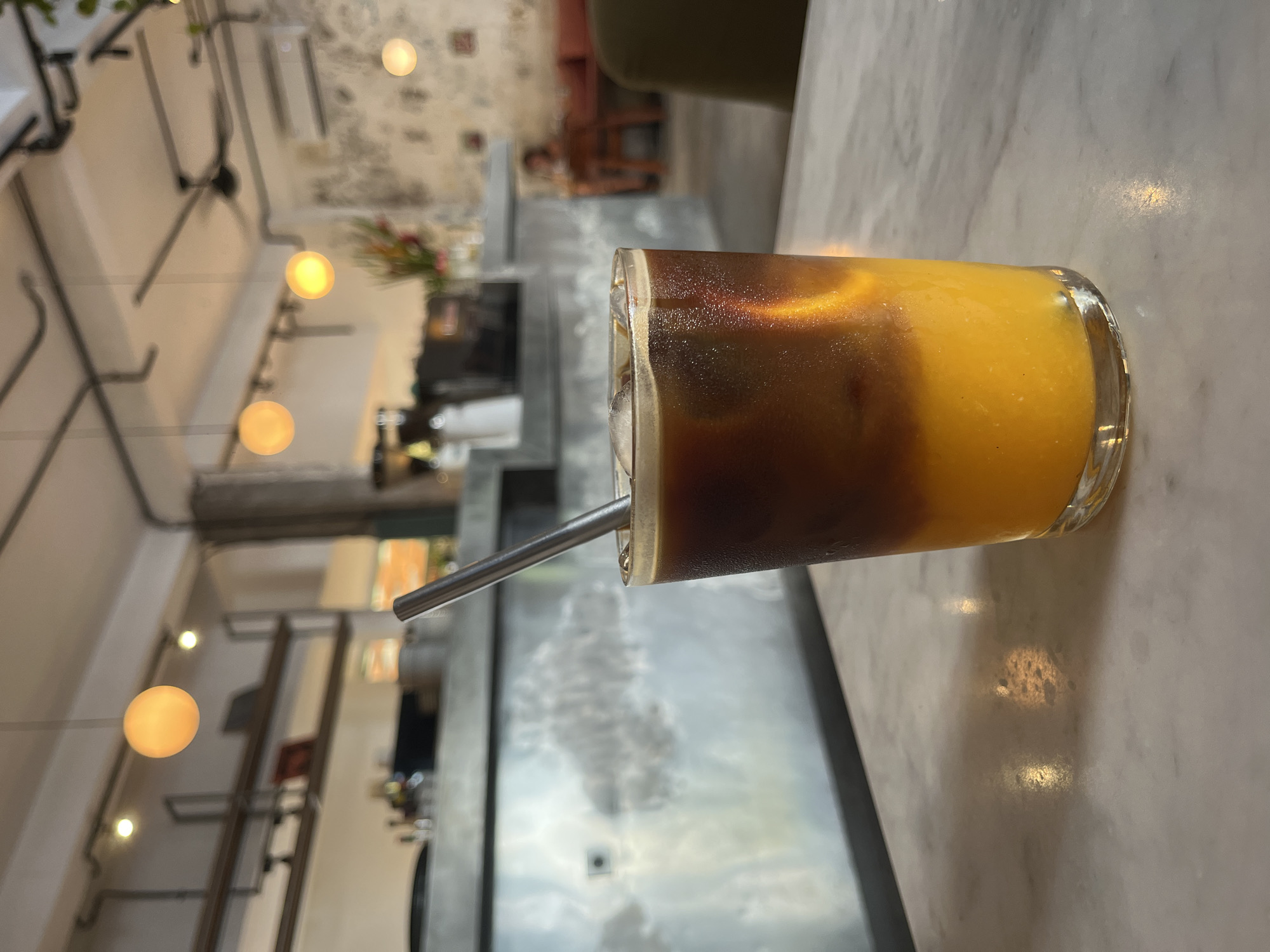 A close-up shot shows a glass filled with a layered drink, with a dark, coffee-colored layer on top and a vibrant orange layer at the bottom, set on a marble table. A silver metal straw is inserted into the drink. The background reveals a blurred interior of a cafe or restaurant with a rustic aesthetic, including exposed pipes, decorative lighting, and textured walls. The location is North Kuta, Indonesia.