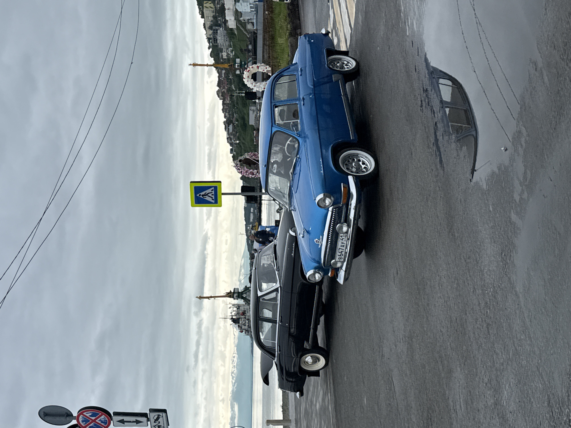 Two vintage GAZ cars, one black and one blue, are parked on a wet asphalt surface in the city of Petropavlovsk-Kamchatsky, Russia. The black car, a GAZ-21 Volga, is parked to the left with its trunk open, revealing its interior. The blue car, also a GAZ-21 Volga, is parked in front of it, facing right. Both cars have chrome accents and white-wall tires.
Behind the cars, the seascape of Avacha Bay is visible, with a cargo ship and mountains in the background. The sky is overcast with dramatic clouds. A pedestrian crossing sign is visible to the left of the blue car, and another traffic sign with a red circle and a diagonal cross, indicating no stopping, is positioned above it. Decorative floral arches are set up behind the cars, suggesting an event or celebration. The wet pavement reflects the cars and the sky, indicating recent rain. The scene appears to be a waterfront area in Petropavlovsk-Kamchatsky, a port city known for its stunning natural scenery and volcanic landscapes.