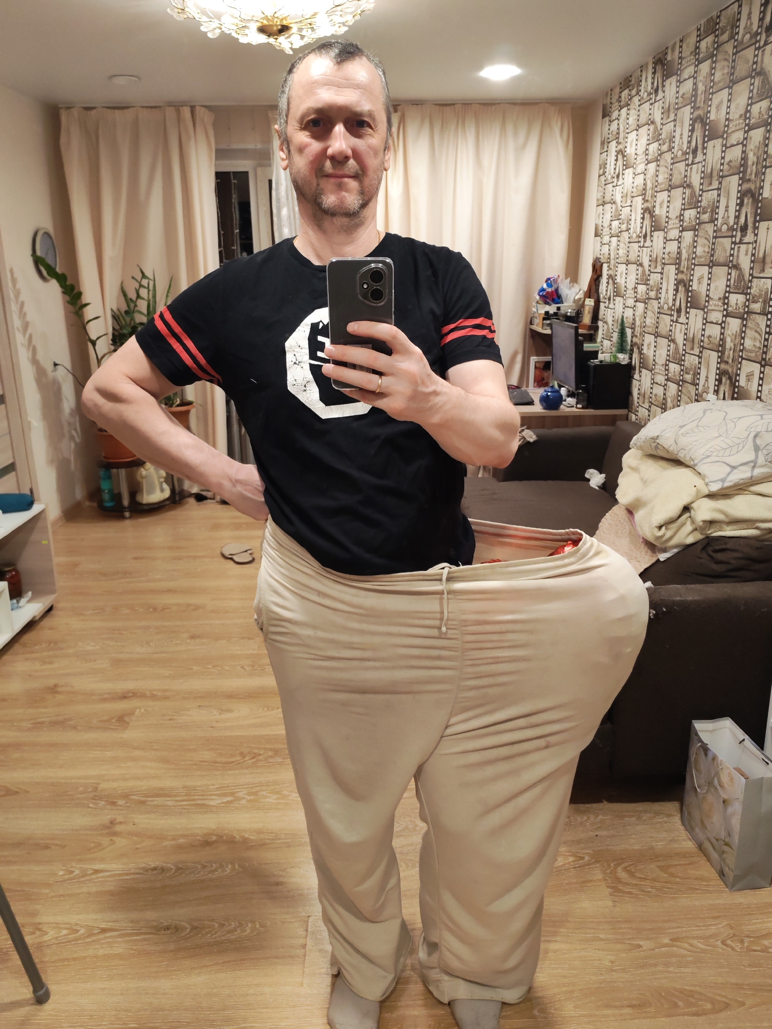 A middle-aged man with short, graying hair is taking a selfie in an interior space, likely a home, located in Chaikovsky, Russia. He wears a black T-shirt with red stripes on the sleeves and a white graphic on the front. He is wearing oversized, beige-colored pants. The background shows a room with light wooden floors, a beige wall with a repetitive film strip design, light-colored curtains, and a dark brown sofa with folded blankets. To the left, a plant sits on a shelf. In the background, a TV, various decorative items, and a gift bag are visible. The lighting is diffused, coming from an overhead ceiling light fixture.