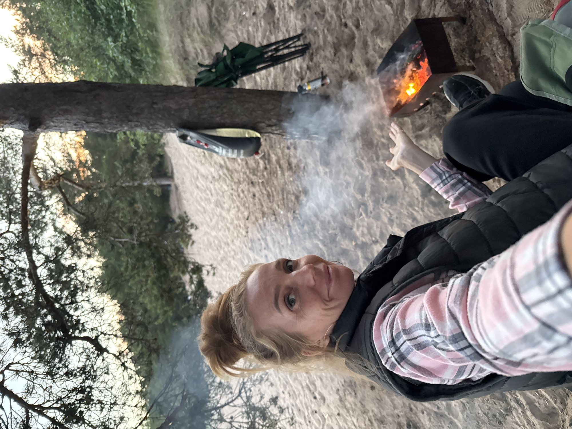 A medium-shot, selfie-style image shows a blonde woman with her hair in a bun, wearing a pink and grey plaid shirt and a black vest, sitting on sandy ground near a campfire. She is looking towards the camera and smiling. To her right, a small black barbecue grill with glowing coals and smoke is visible. Behind her, a large tree stands with camping chairs and other equipment leaning against it. The background consists of sand dunes and sparse pine trees, suggesting an outdoor, possibly coastal, environment. The overall lighting indicates it is either dawn or dusk. The scene is set in the city of Zelenaya Roshcha, Russia.